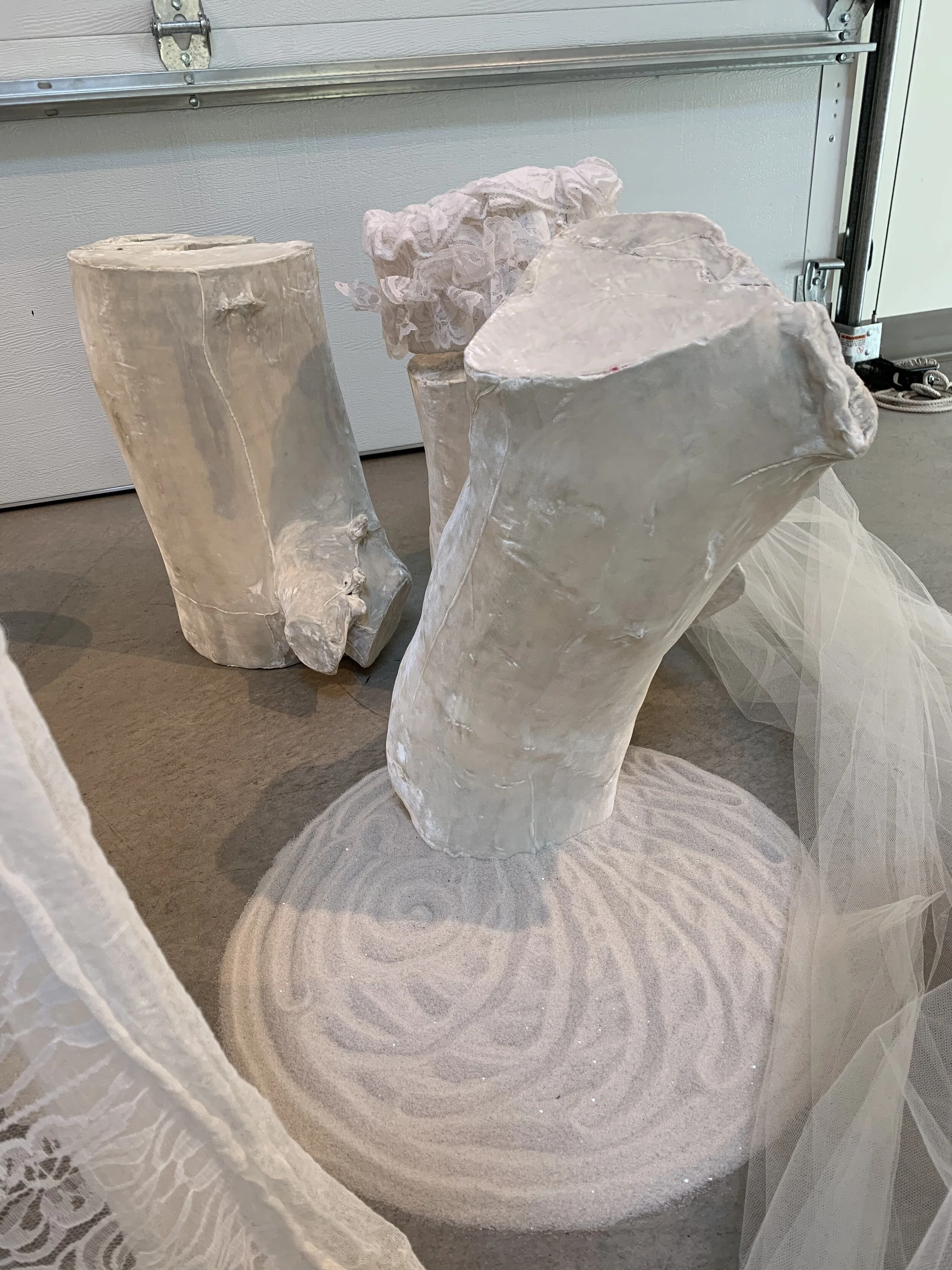Three large white mannequin legs made of a material resembling plaster or fiberglass, positioned on a circular base with decorative patterns, with lace fabric draped around them, in a garage or workshop setting.