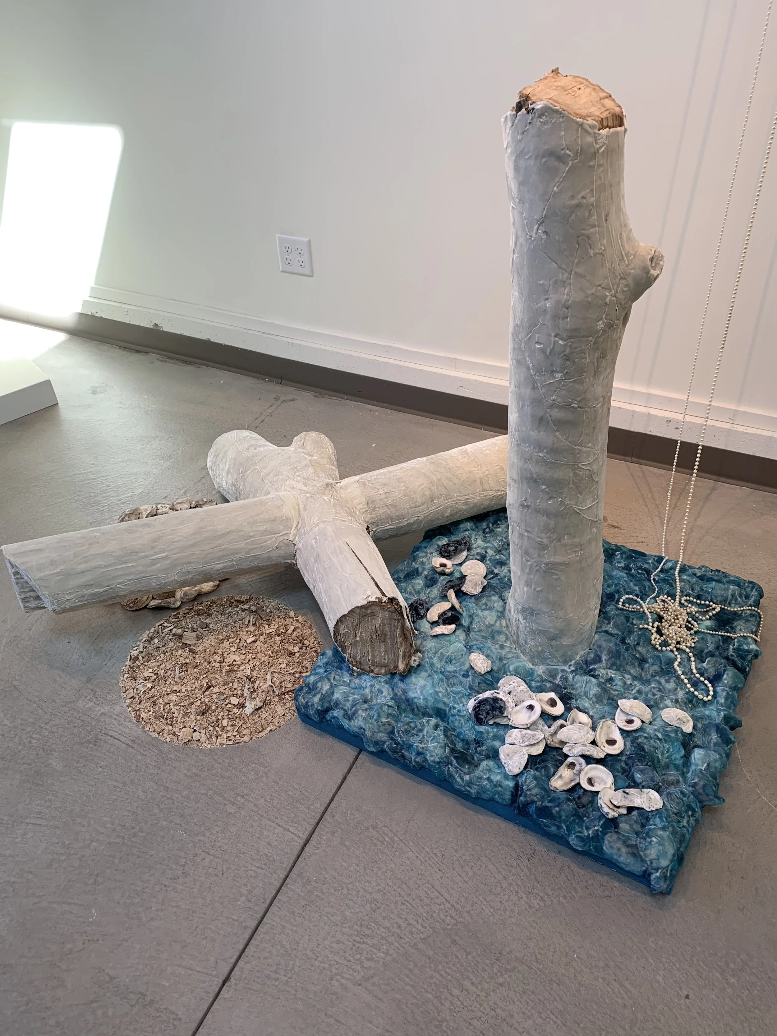 A sculpture of a tree, cross, and various natural elements arranged on the floor, with seashells and a pearl necklace on a blue textured base.