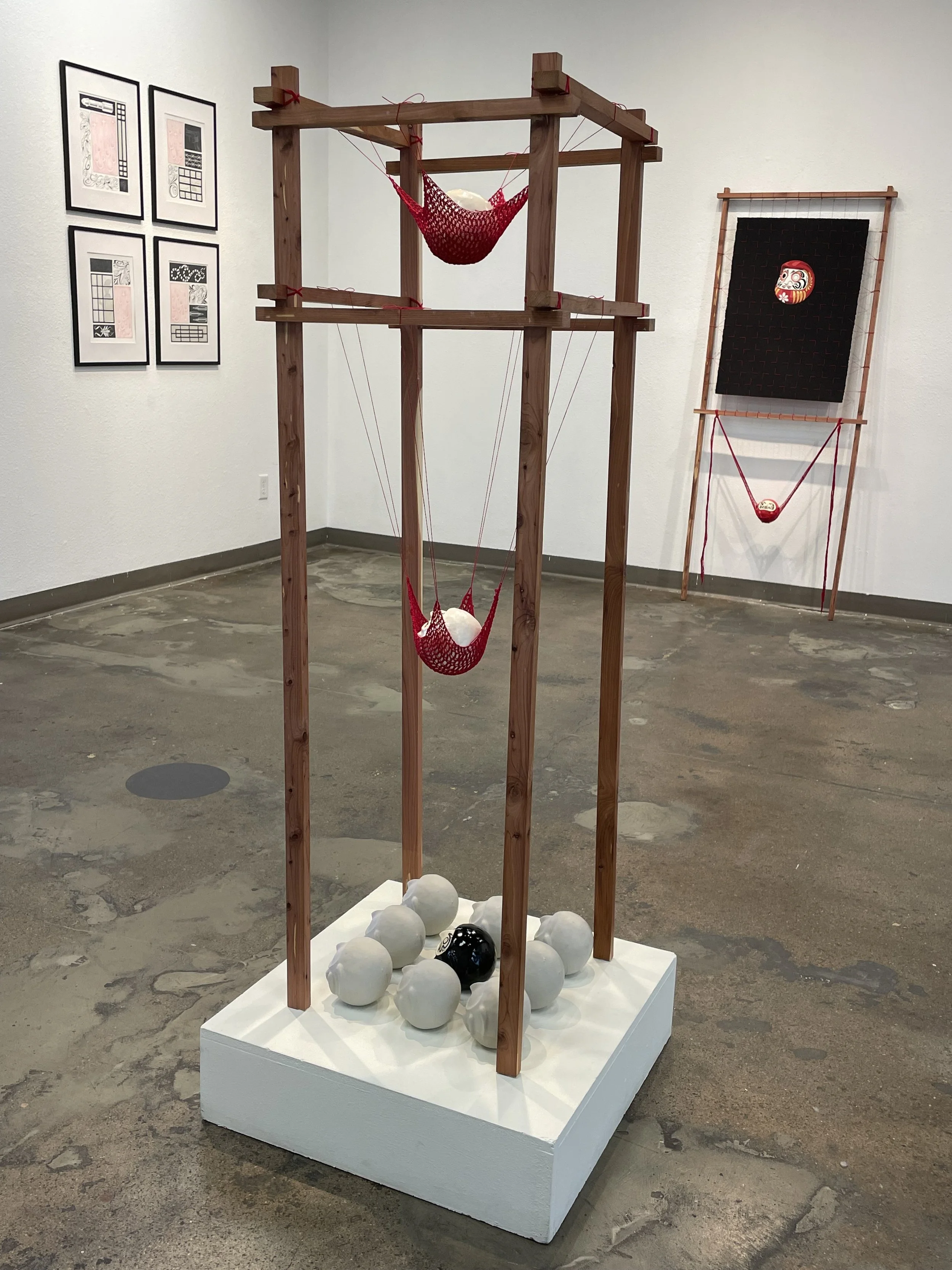 Art installation with two red hammocks suspended from a wooden framework, surrounded by white and black spheres on a white platform. The background includes framed abstract art on the wall and another hanging art piece with a black fabric and a red a