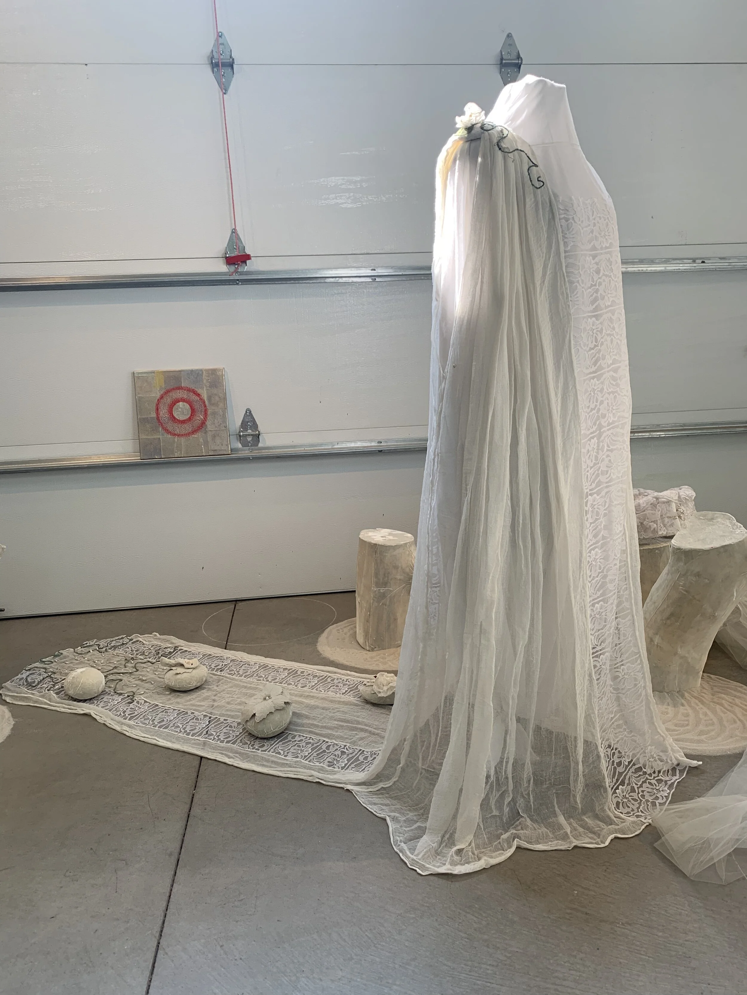 Display of white lace and sheer fabric on mannequins, surrounding wooden stumps, decorative stones, and fabric with lace patterns on the floor, against a garage door background.