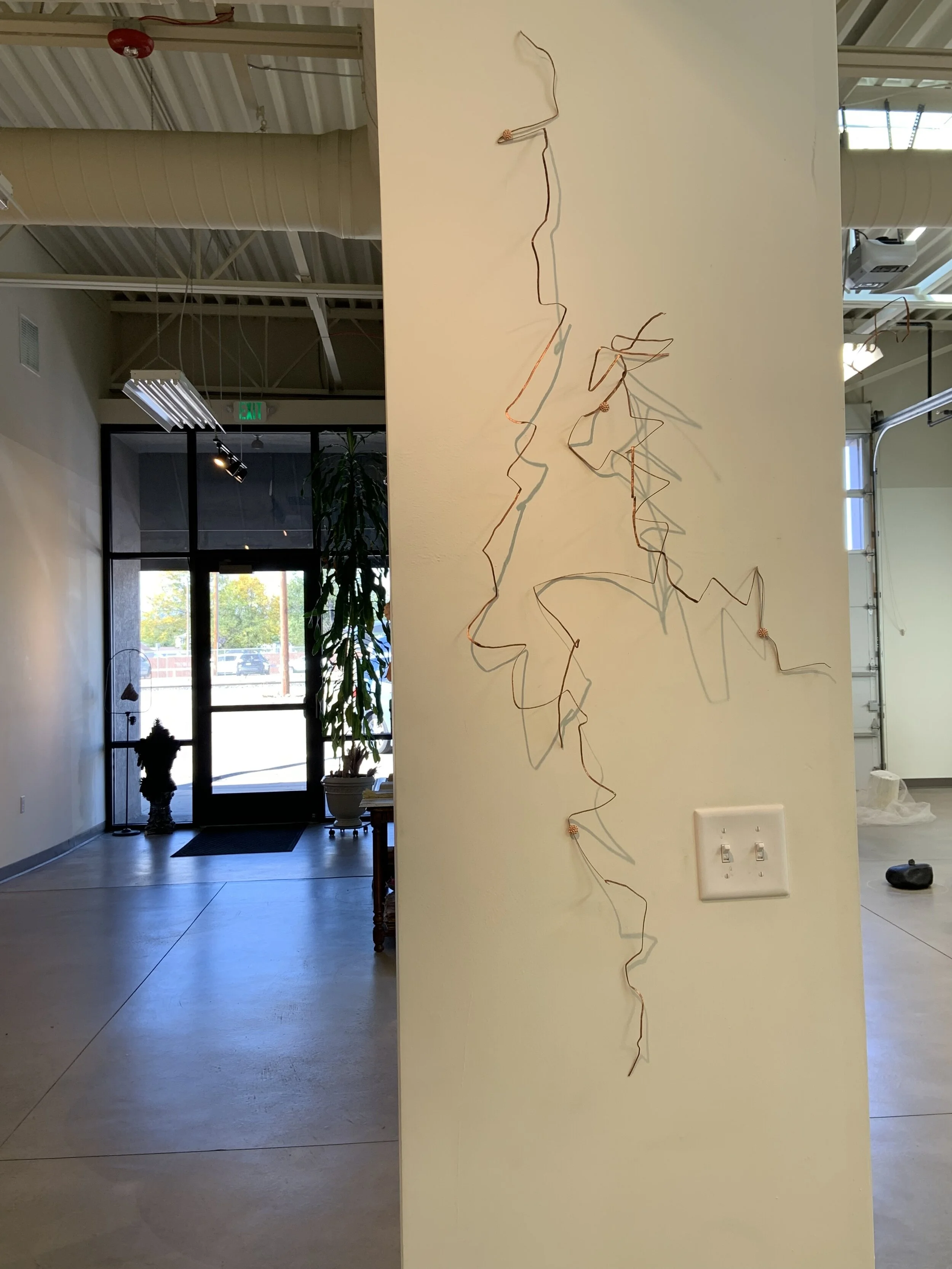 Decorative wire art hanging on a beige wall in an industrial-style interior with a glass door, plants, and profile of a dog in the background.