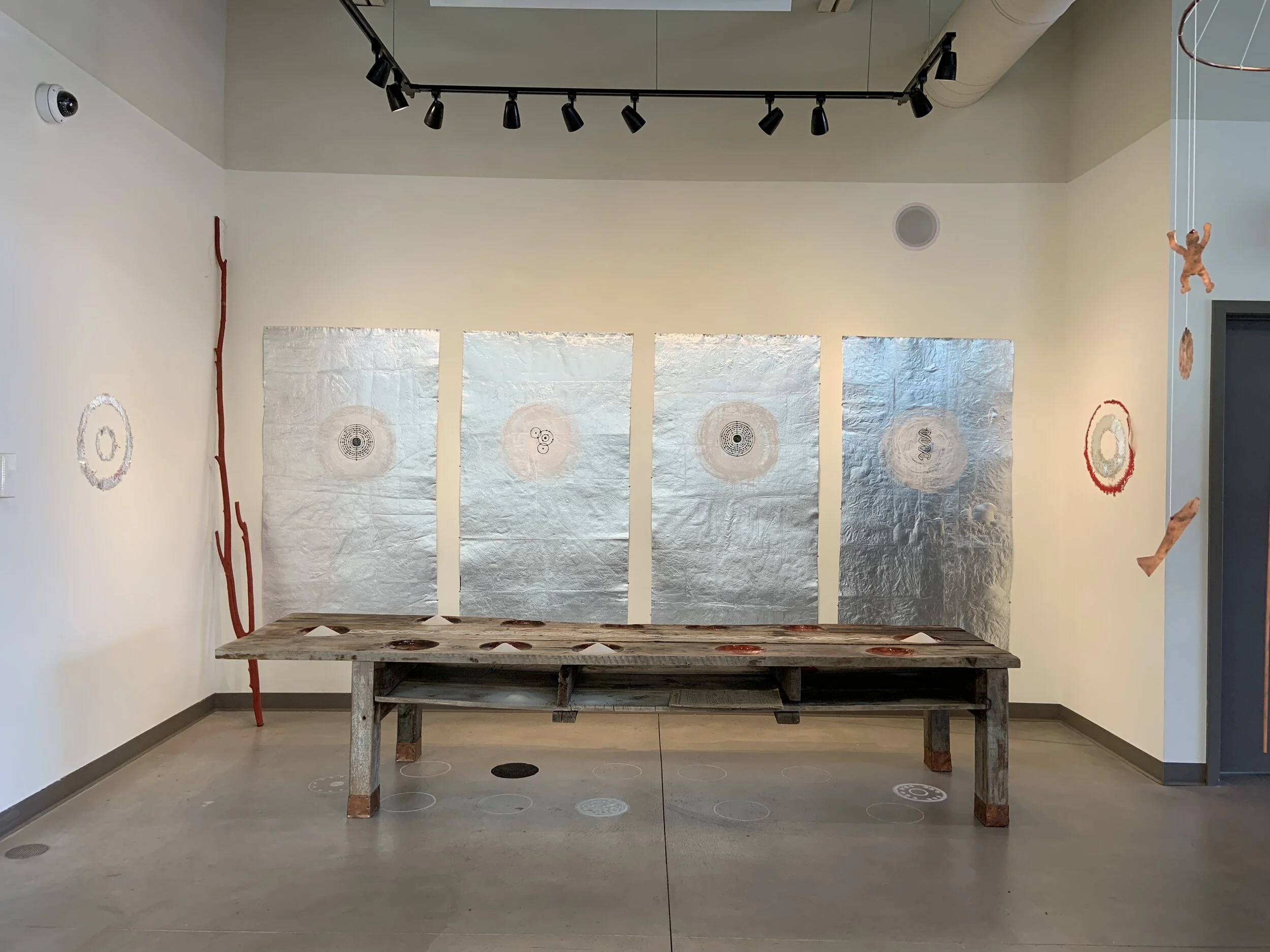Contemporary art installation with four metallic panels, circular symbols, and various paper and string sculptures in a gallery with track lighting and a wooden bench.