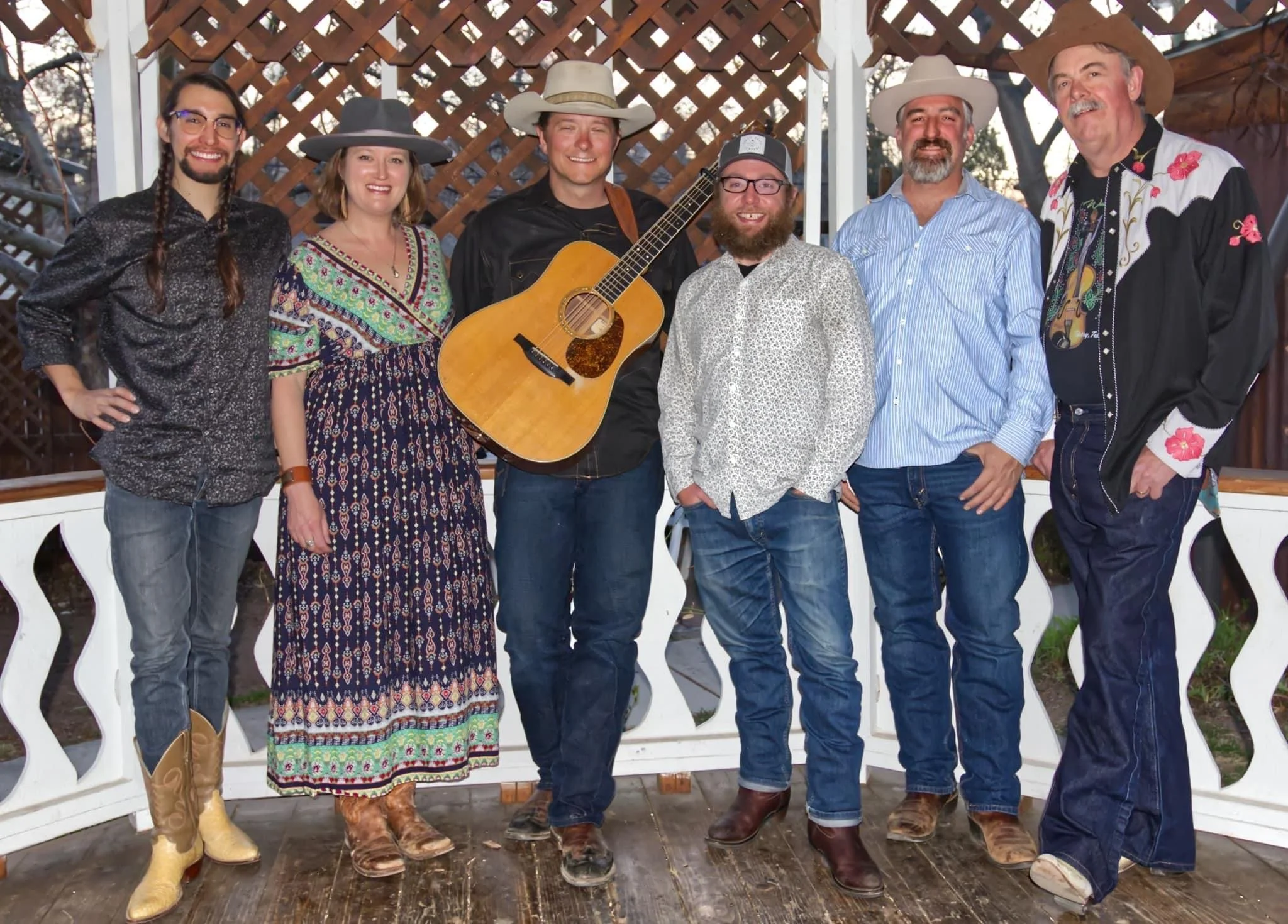 Group of seven people, four men and three women, standing outdoors, some wearing cowboy hats, with one holding a guitar, in front of a wooden lattice backdrop.