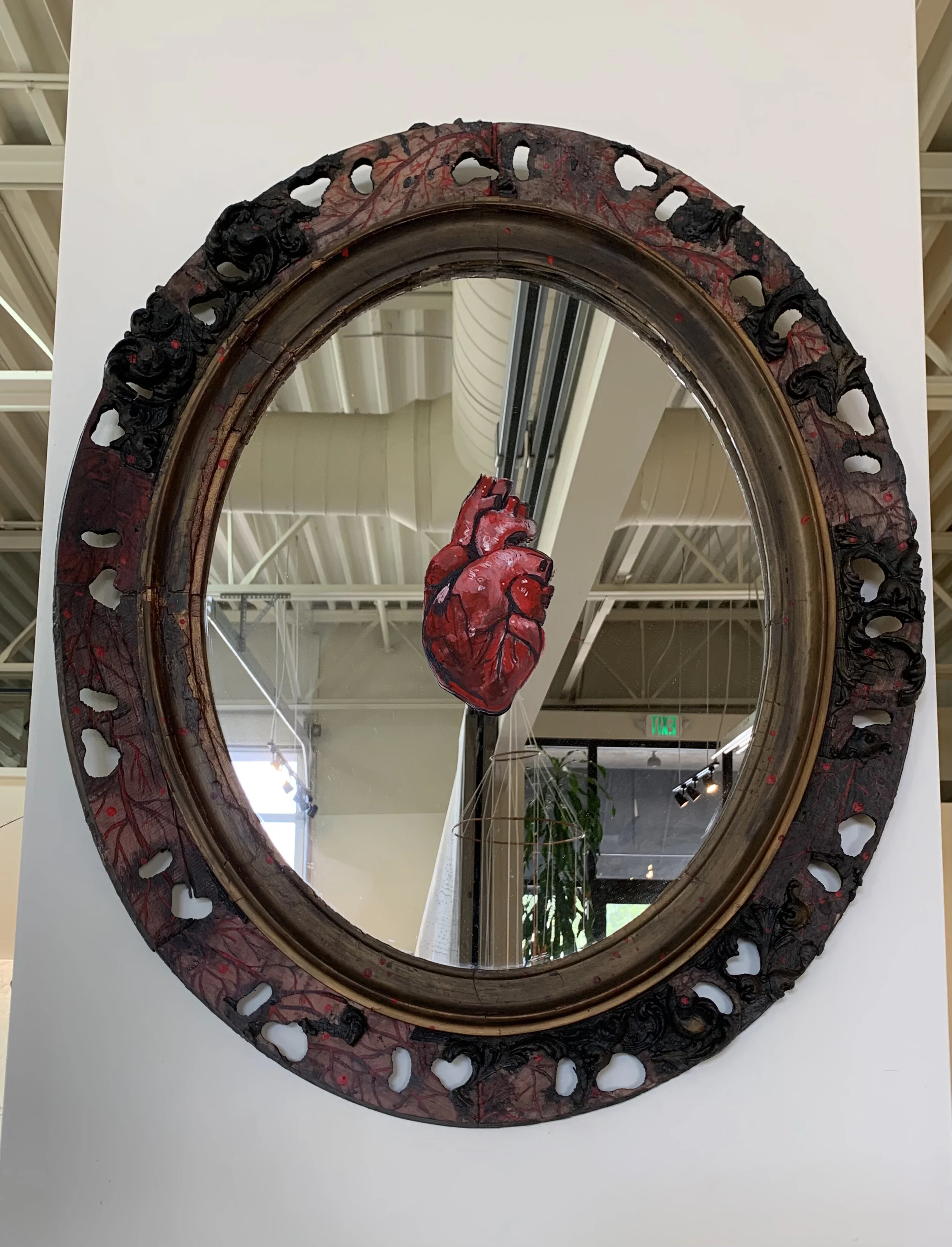 Decorative mirror with a black and red ornate rim, featuring a red anatomical heart sculpture in the reflection.