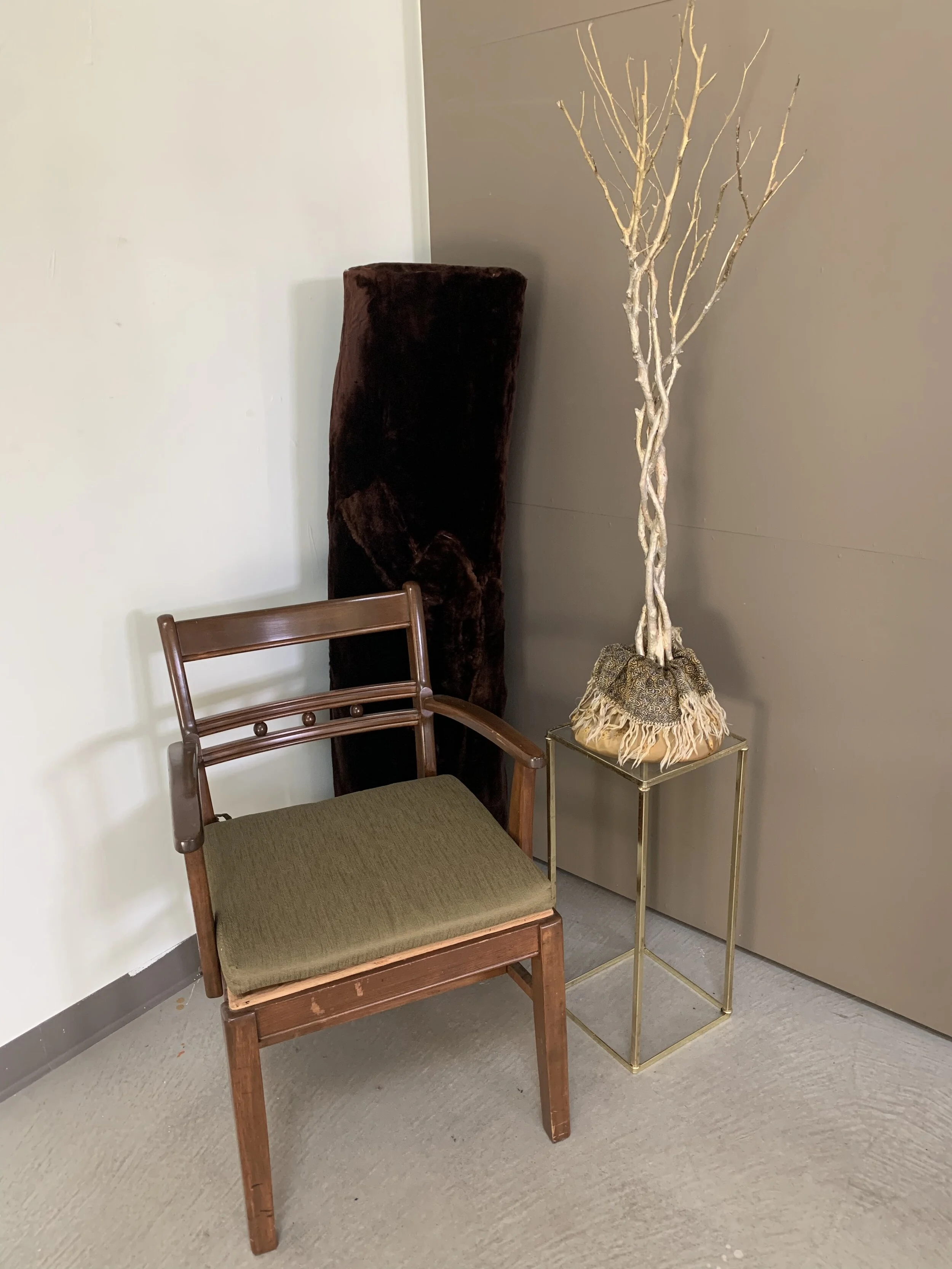 An antique wooden chair with a green cushion, a tall dried bare tree in a decorative container, and a tall dark brown padded furniture piece in the corner.