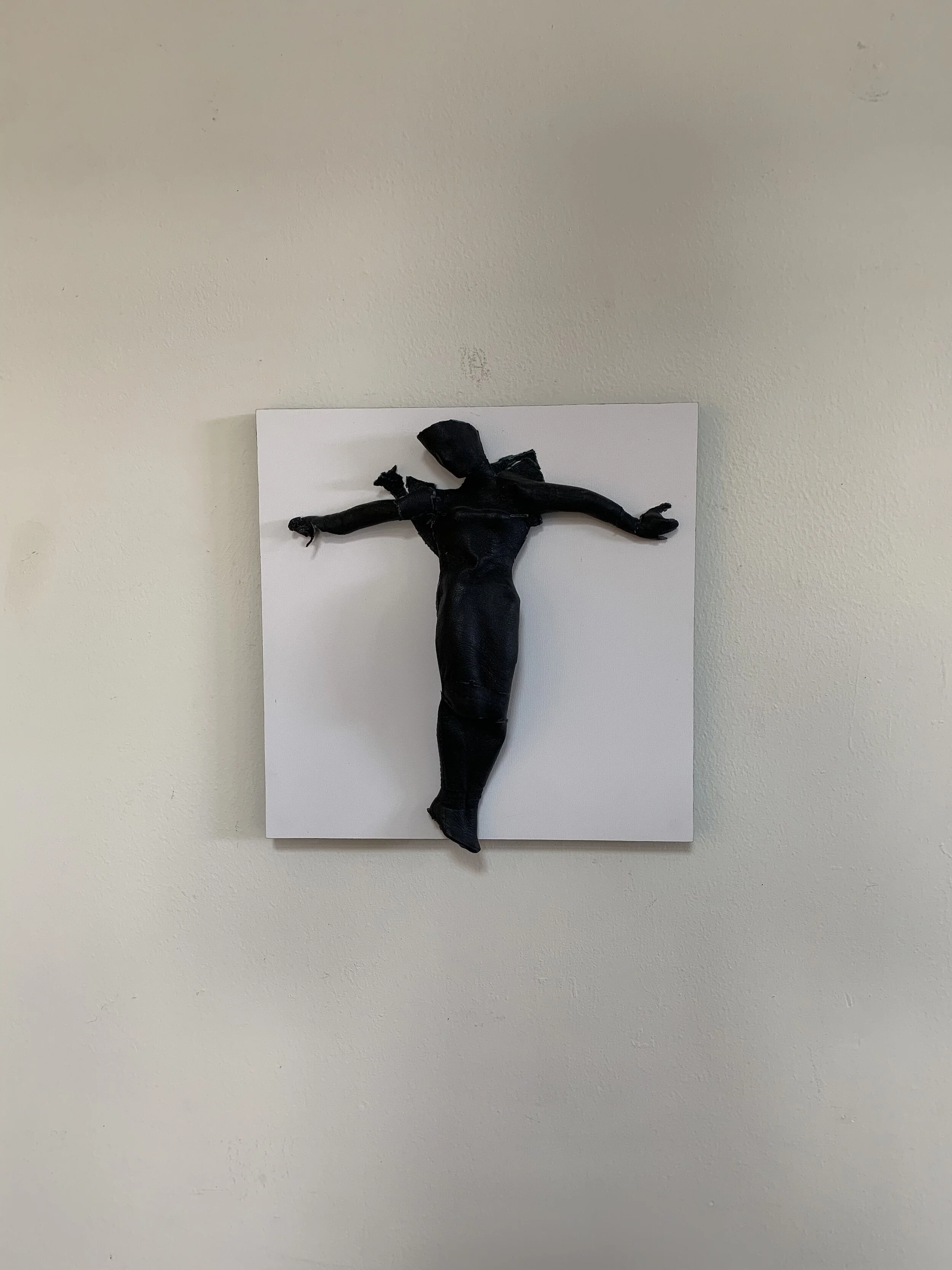 A wall sculpture resembling a human figure with arms outstretched, made from black material, mounted on a white square panel on a light-colored wall.