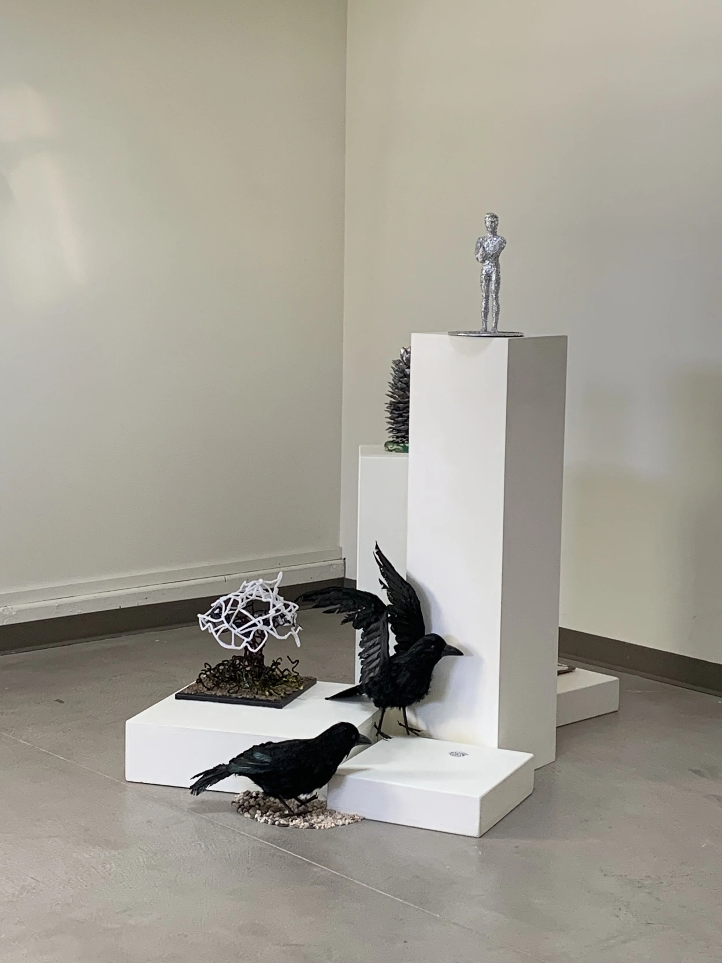 Sculpture display with a silver human figure on a white pillar, black bird sculptures, a white corncob sculpture, and abstract shapes on a gray tiled floor.