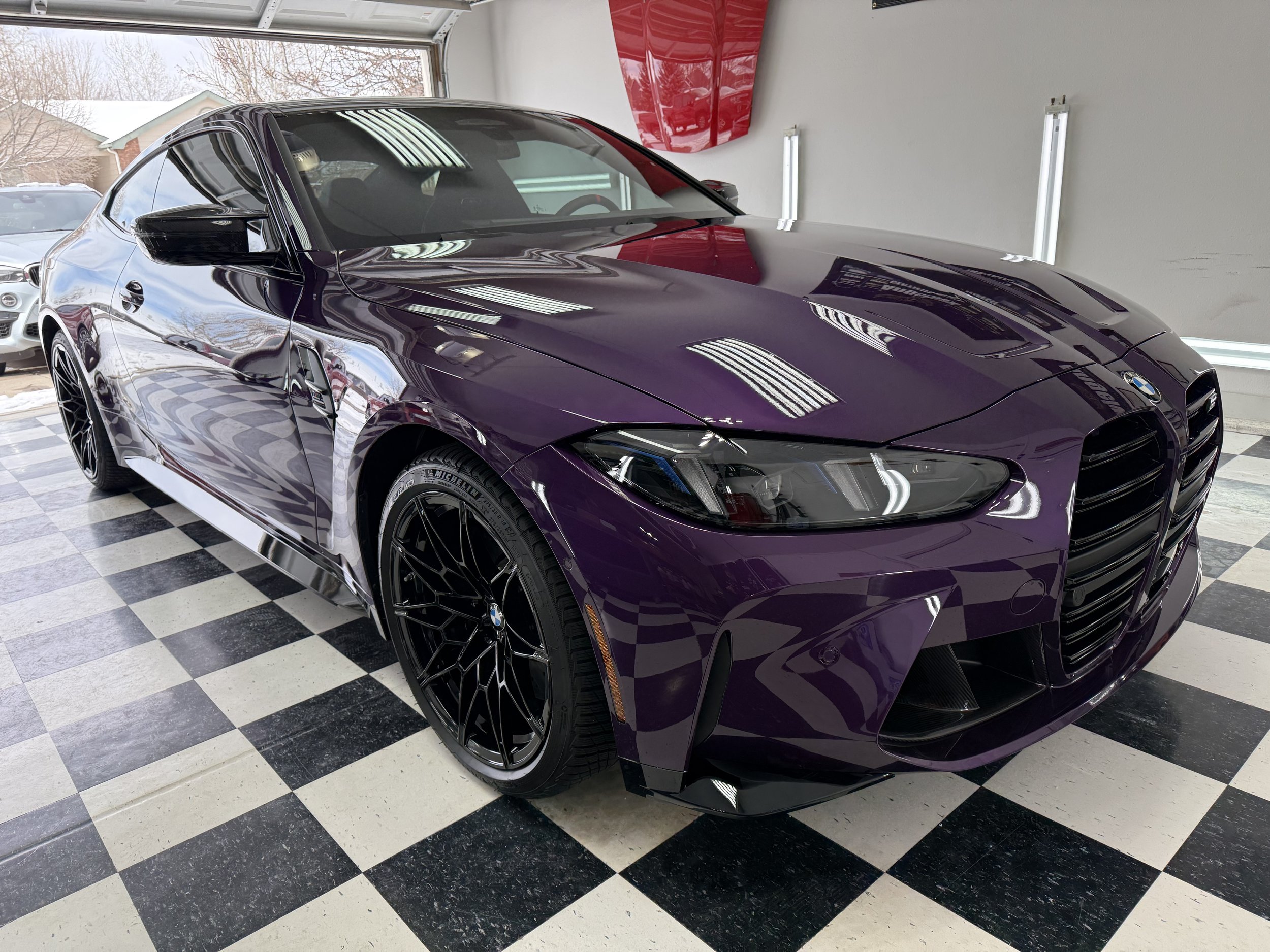 A purple BMW sports car parked inside a showroom with black and white checkered floors, reflecting ceiling lights and windows in the shiny surface of the car.