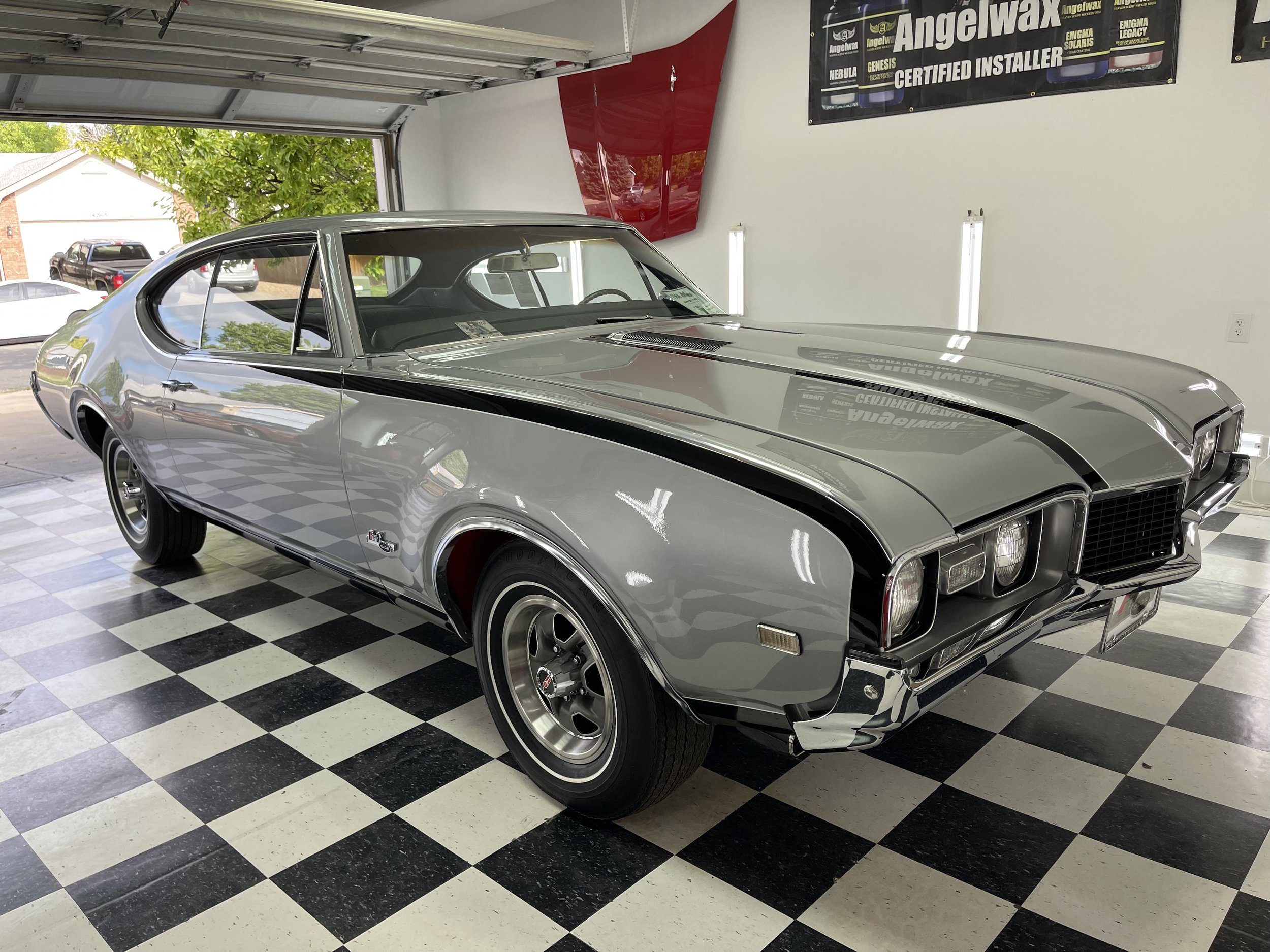 A vintage silver Chevrolet car parked inside a garage with a black and white checkered floor. The garage has an open door revealing parked cars outside and a green tree. There is a banner on the wall advertising Angelwax products.