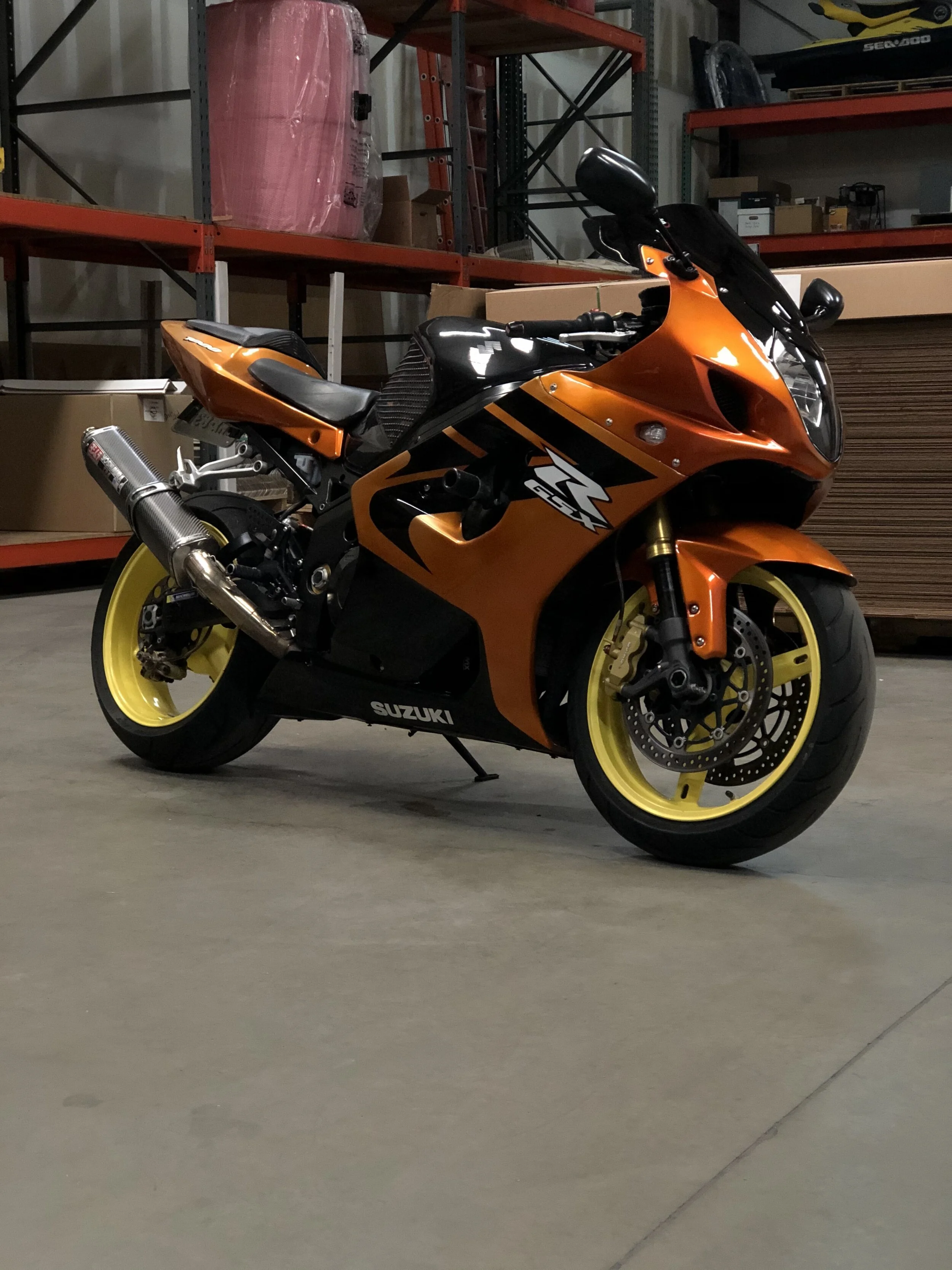An orange and black Suzuki sport motorcycle with yellow wheels parked in a warehouse, with shelves in the background holding boxes, a wrapped item, and a kayak.