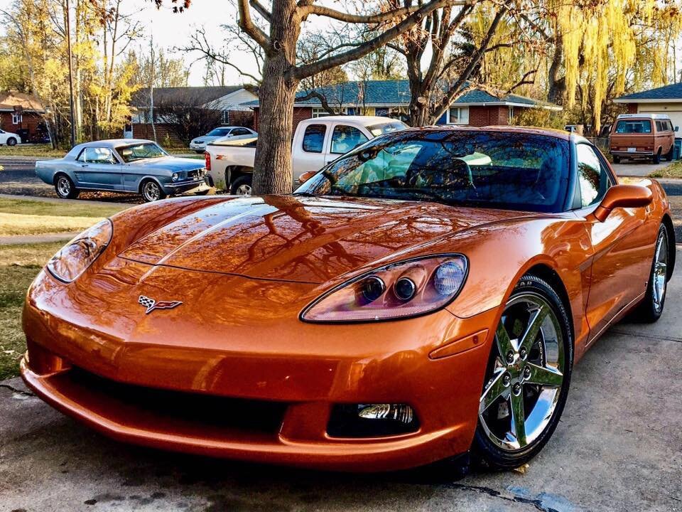 An orange Chevrolet Corvette sports car parked outdoors with a suburban neighborhood and other classic cars in the background.