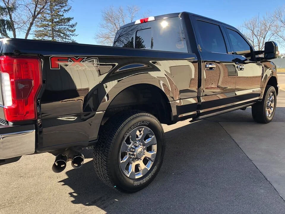 Black Ford F-150 pickup truck with chrome wheels and dual exhaust pipes, parked on a paved driveway during daylight.