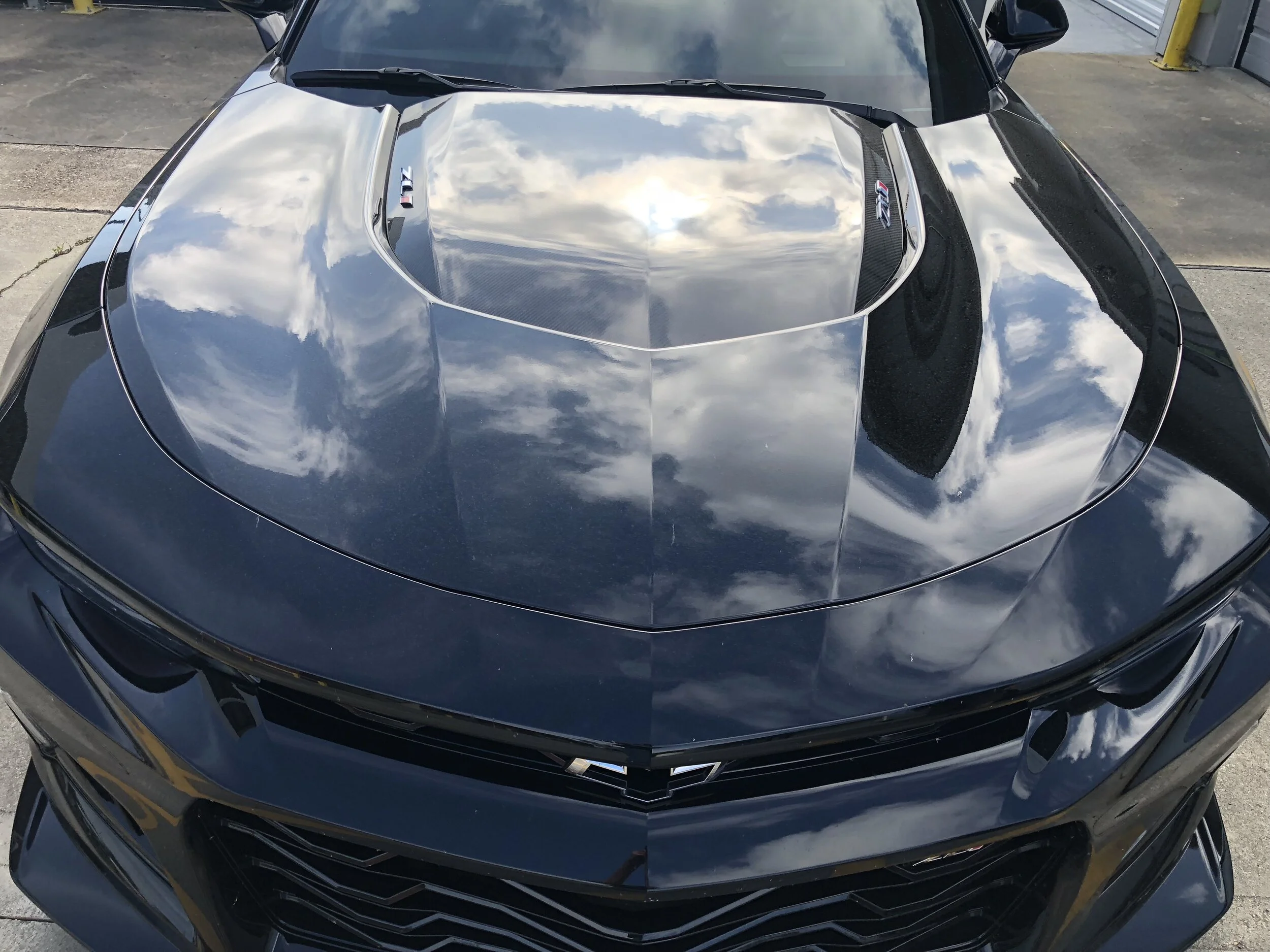 Front view of a black sports car with a reflective hood, displaying a mirror-like reflection of the sky with clouds. The car has a Chevrolet LTZ badge on the hood.