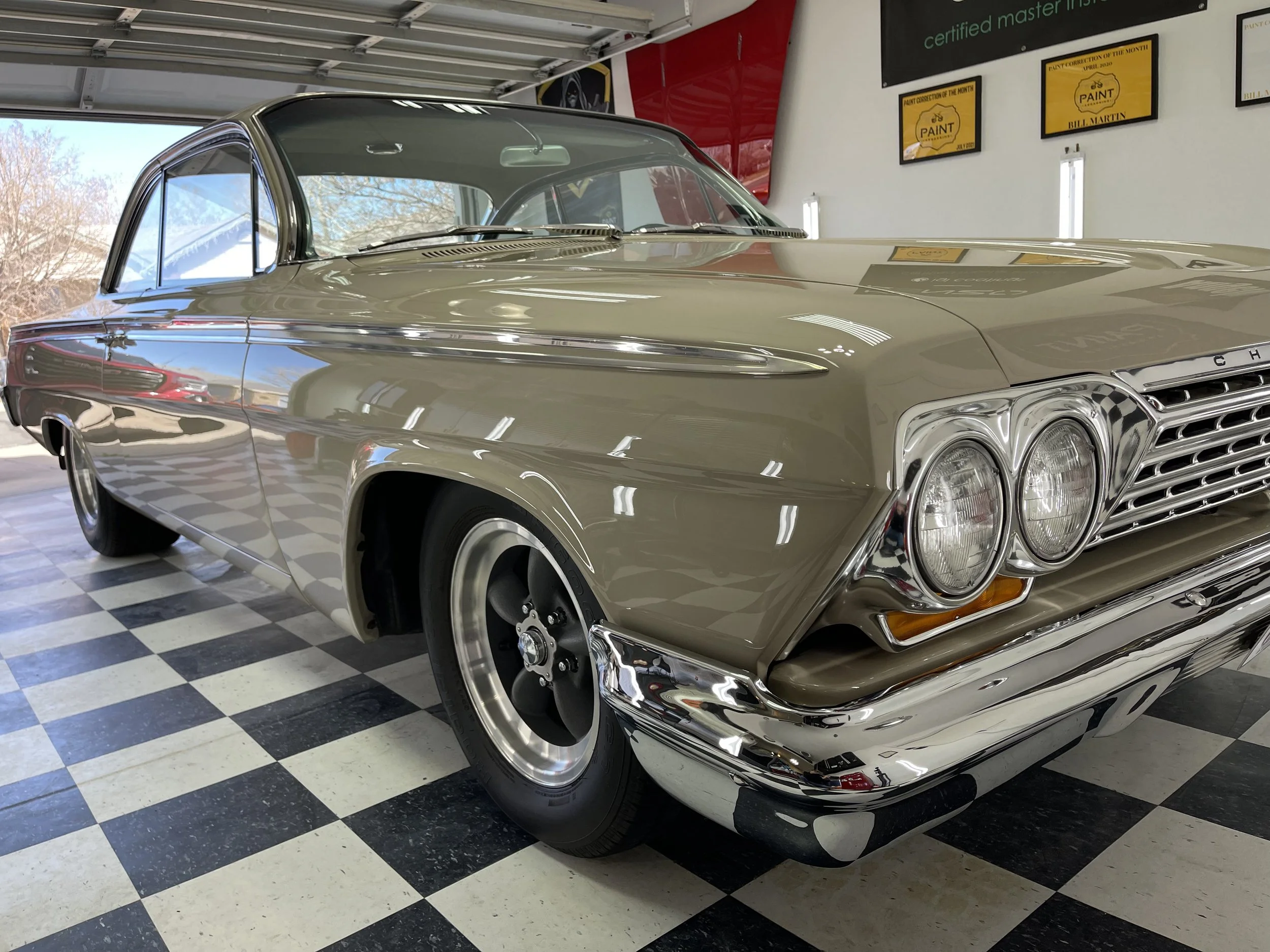 A vintage beige Chevrolet car with a shiny chrome grille and headlight area, parked inside a garage with checkered black and white floor tiles. The garage has posters and plaques on the wall and sunlight is coming through an open garage door.