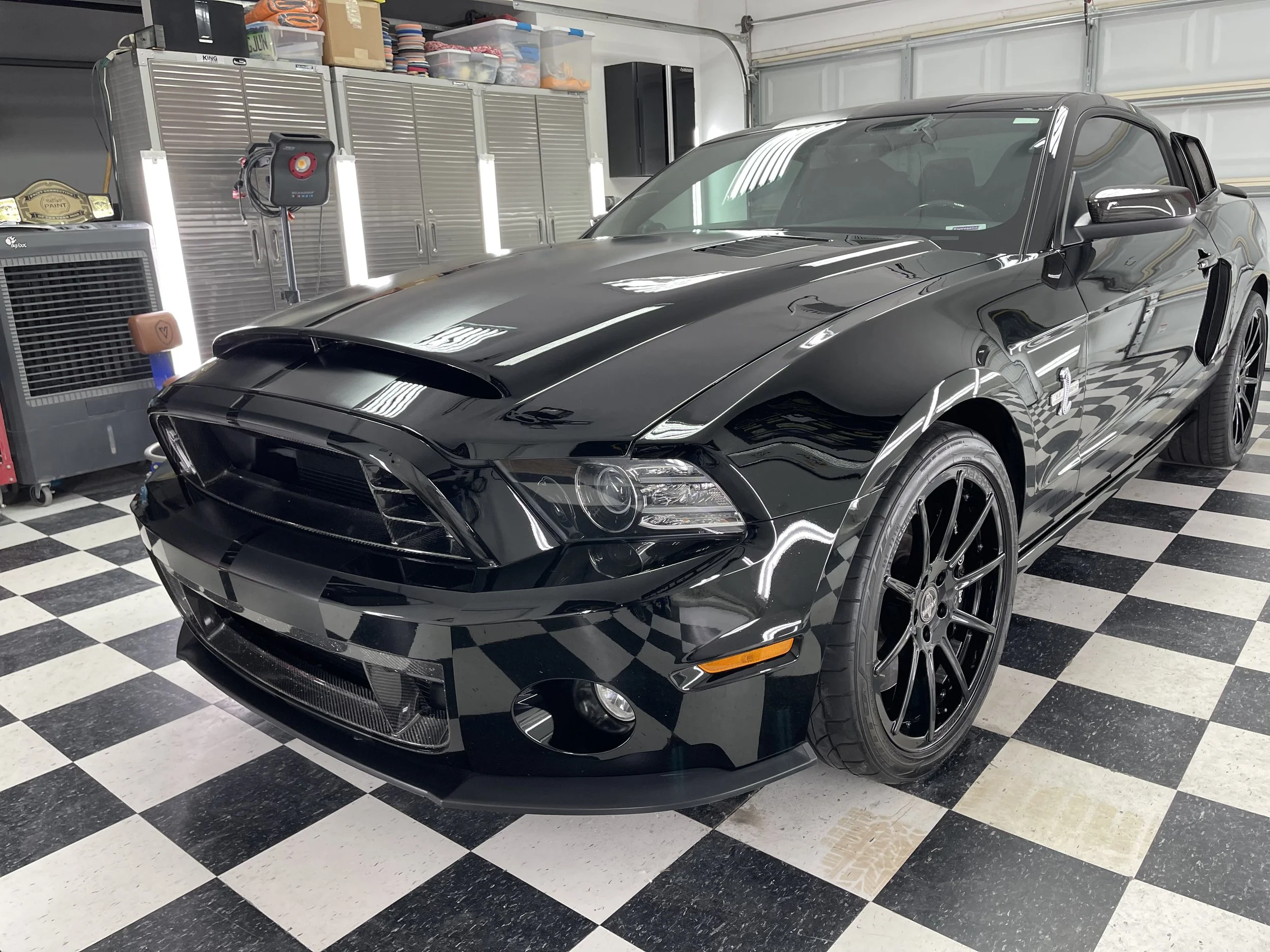 A black sports car parked in a garage with a checkered black and white floor. The garage has storage cabinets, a toolbox, and various items on shelves.