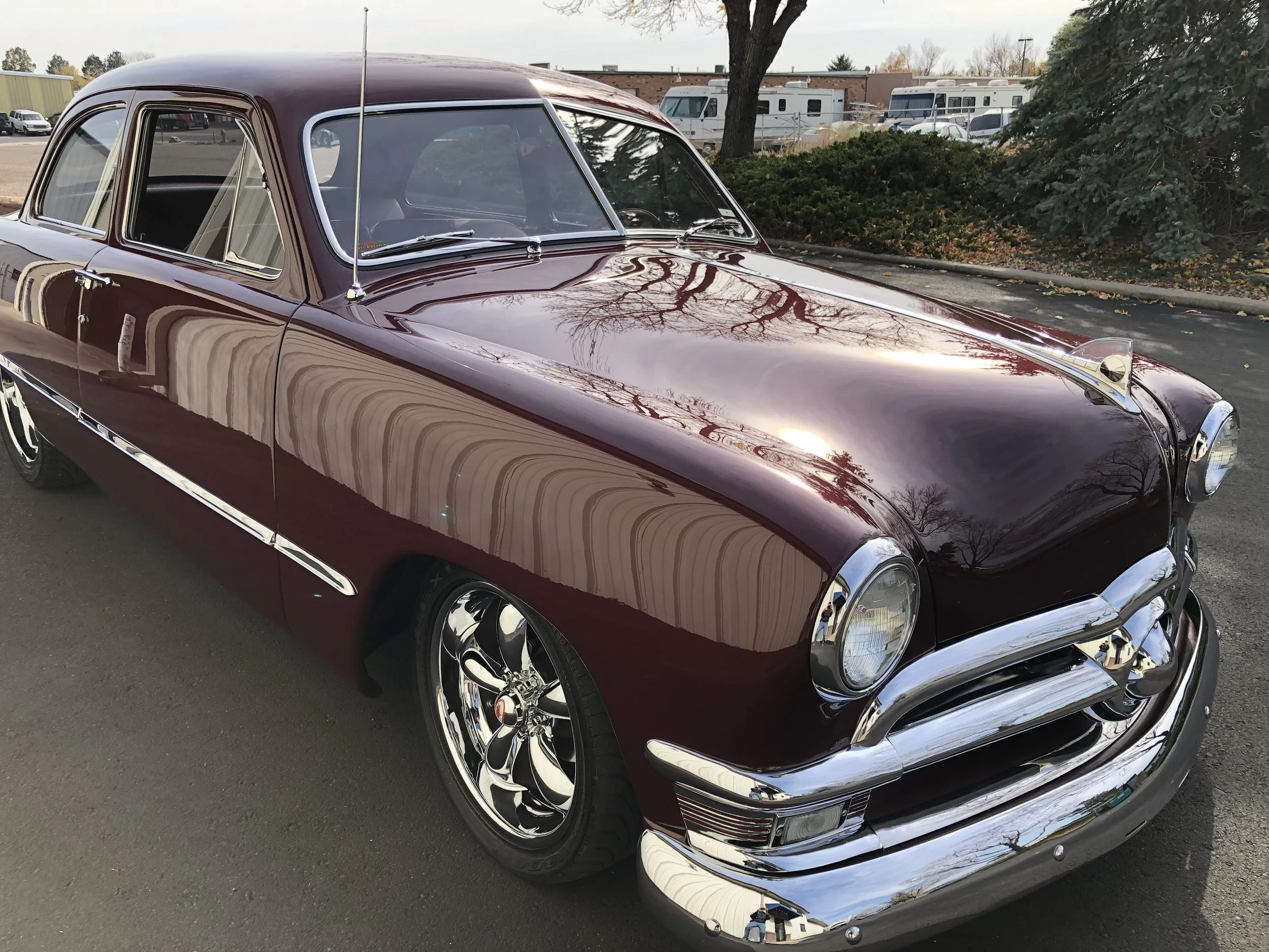 A vintage maroon car parked outdoors on a paved surface, with shiny chrome wheels and trim, reflecting the bare trees surrounding it.