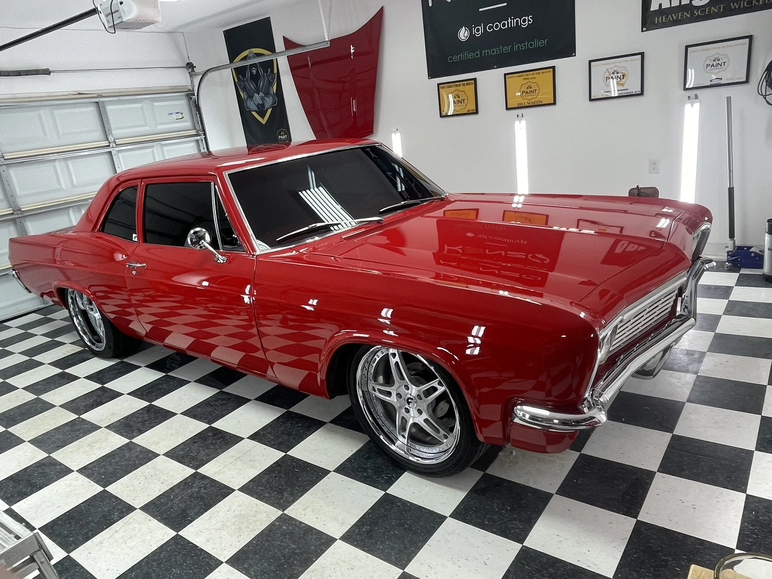 A red vintage muscle car with chrome rims parked inside a garage with checkered black and white floor tiles. The garage has posters and banners on the wall, and a garage door in the background.