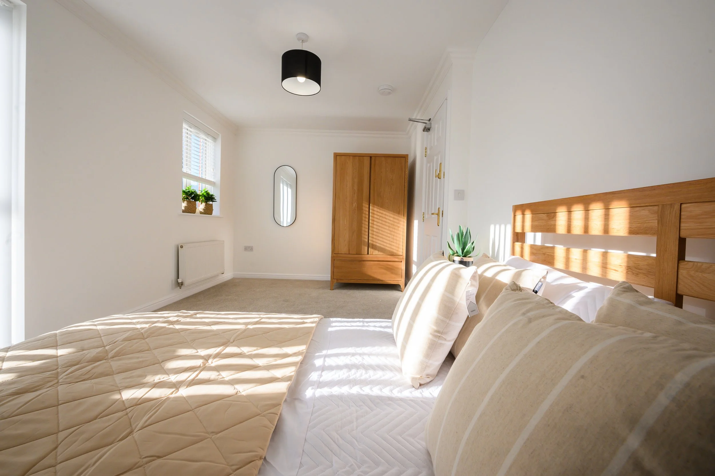 A bright and minimalistic bedroom with a bed featuring beige pillows, a beige quilt, and a wooden headboard. There is a small plant on the bedside table, white walls, a wooden wardrobe, a black ceiling light, a window with blinds, and a mirror on the wall.