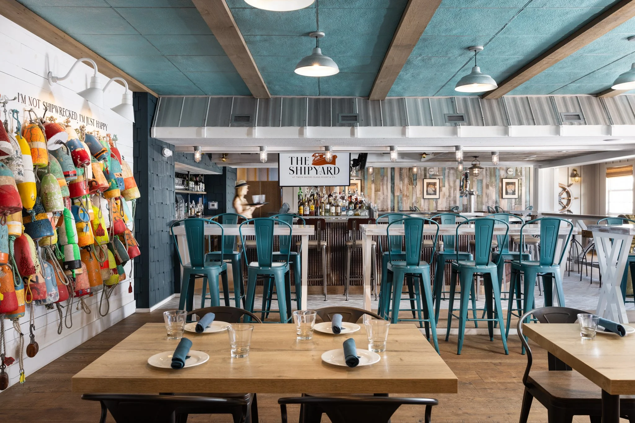 Interior of a restaurant or bar, featuring a decor with a nautical theme, a bar with various alcohol bottles, high teal metal chairs, a wall with colorful buoy decorations, and a sign that reads "The Shipyard".