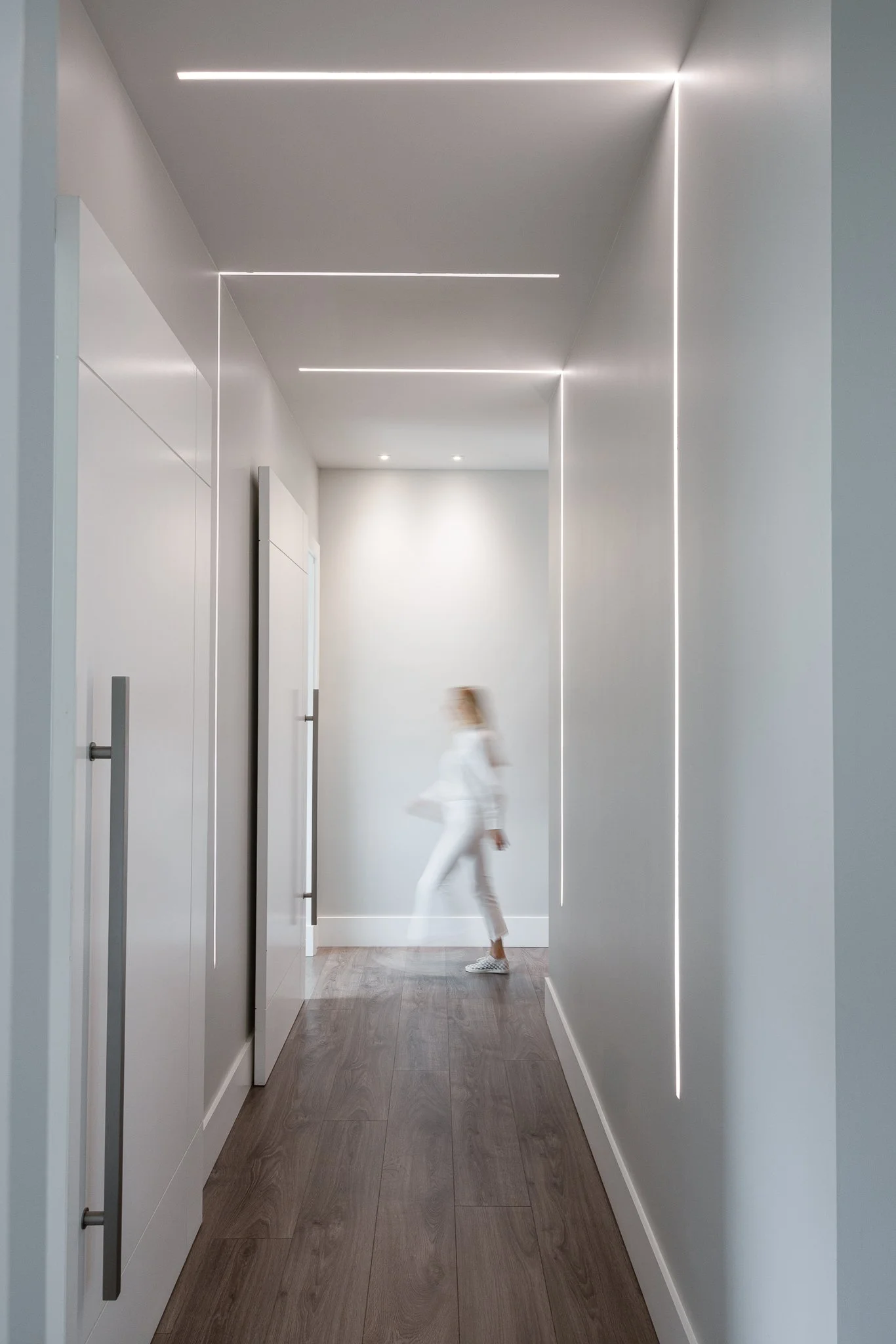 A woman in white walking down a modern, bright hallway with minimalist ceiling lighting and wooden flooring.