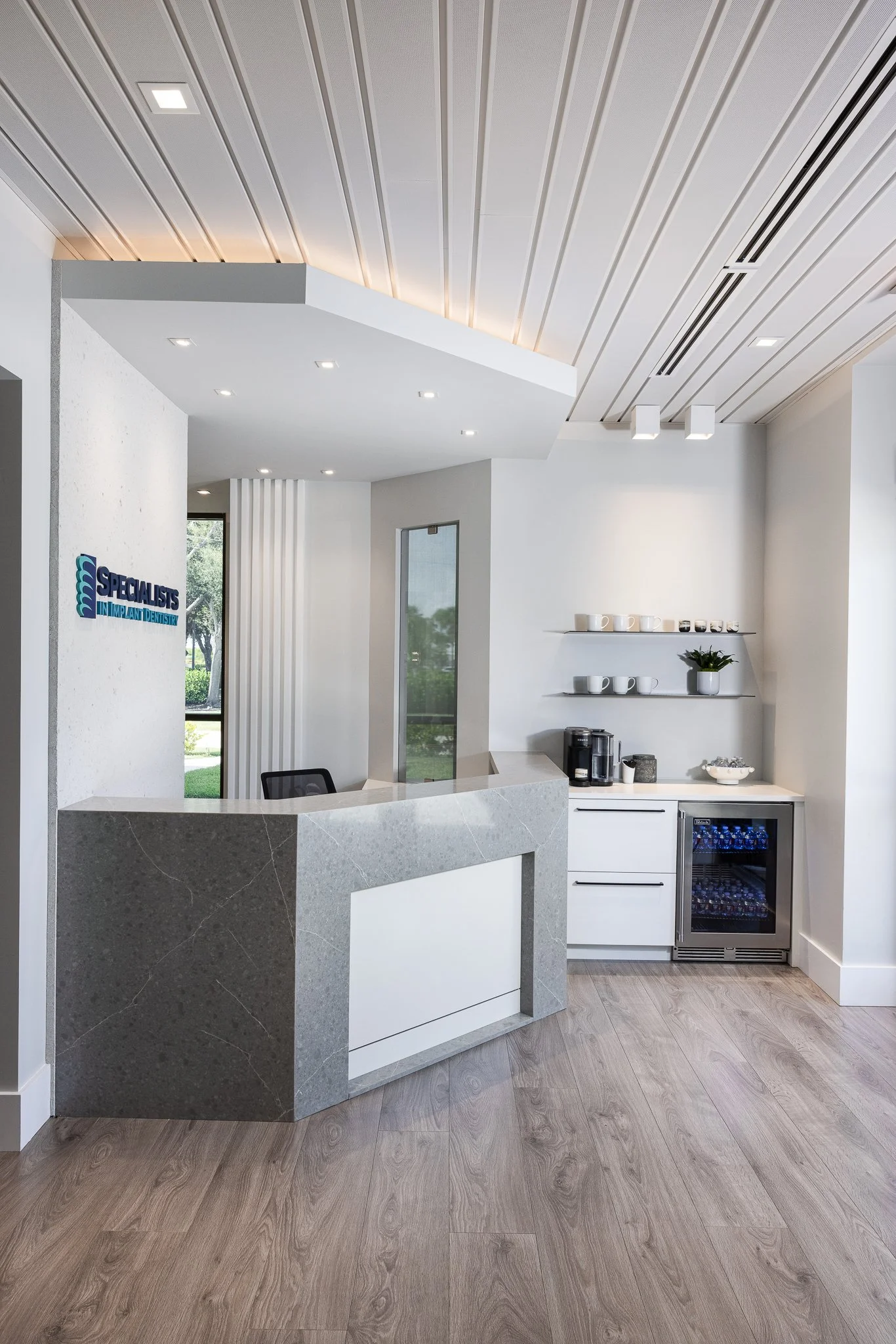 Modern reception area with a stone front desk, white walls, and ceiling with built-in lighting. To the right, there is a small kitchenette with coffee-making equipment, cups, and a mini fridge. A window and glass door are visible in the background, w