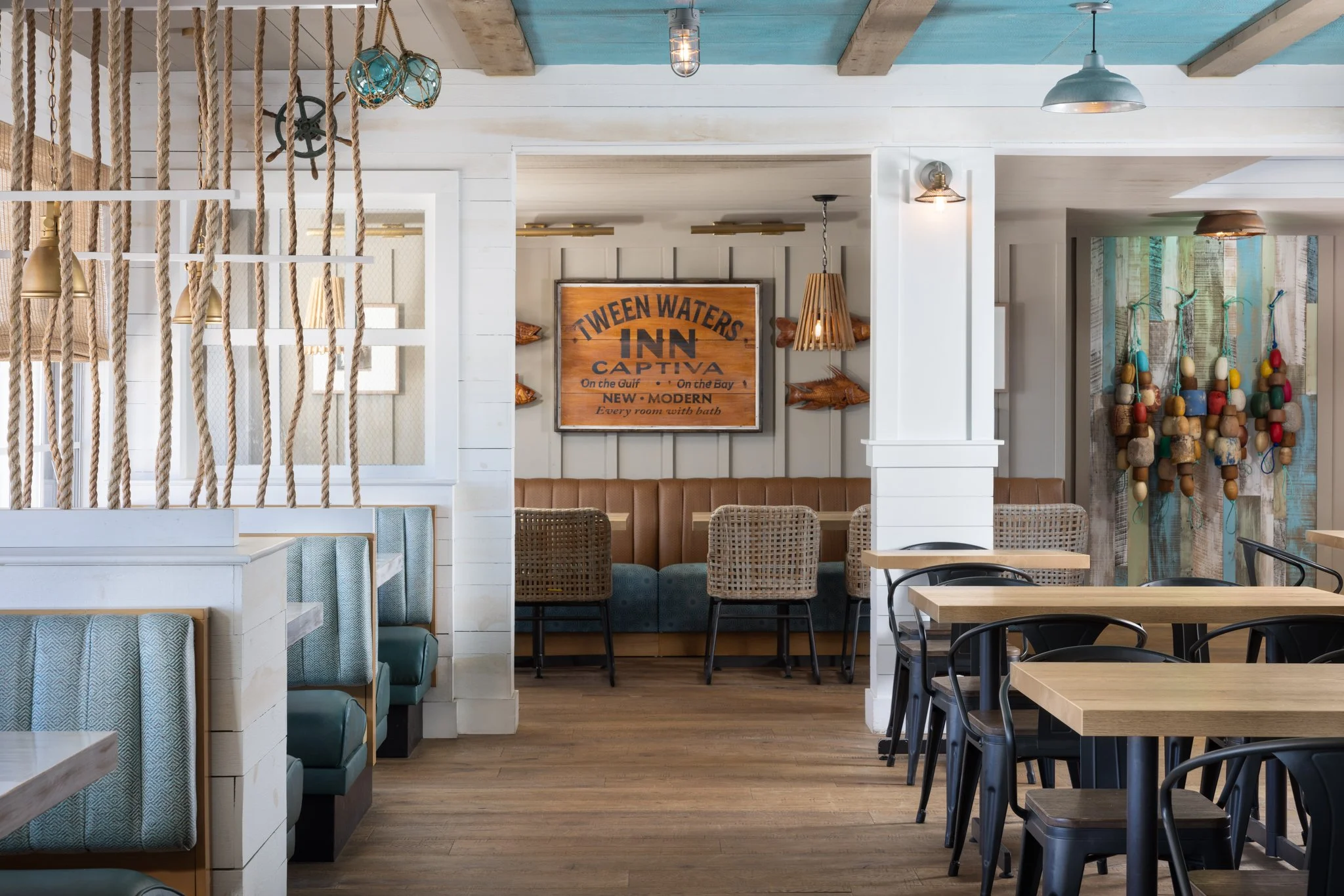 Interior of a seaside restaurant with wooden tables, black and woven chairs, nautical decor, a sign that reads 'TWEEN WATERS INN CAPTIVA,' and colorful buoys and ropes hanging on the wall.