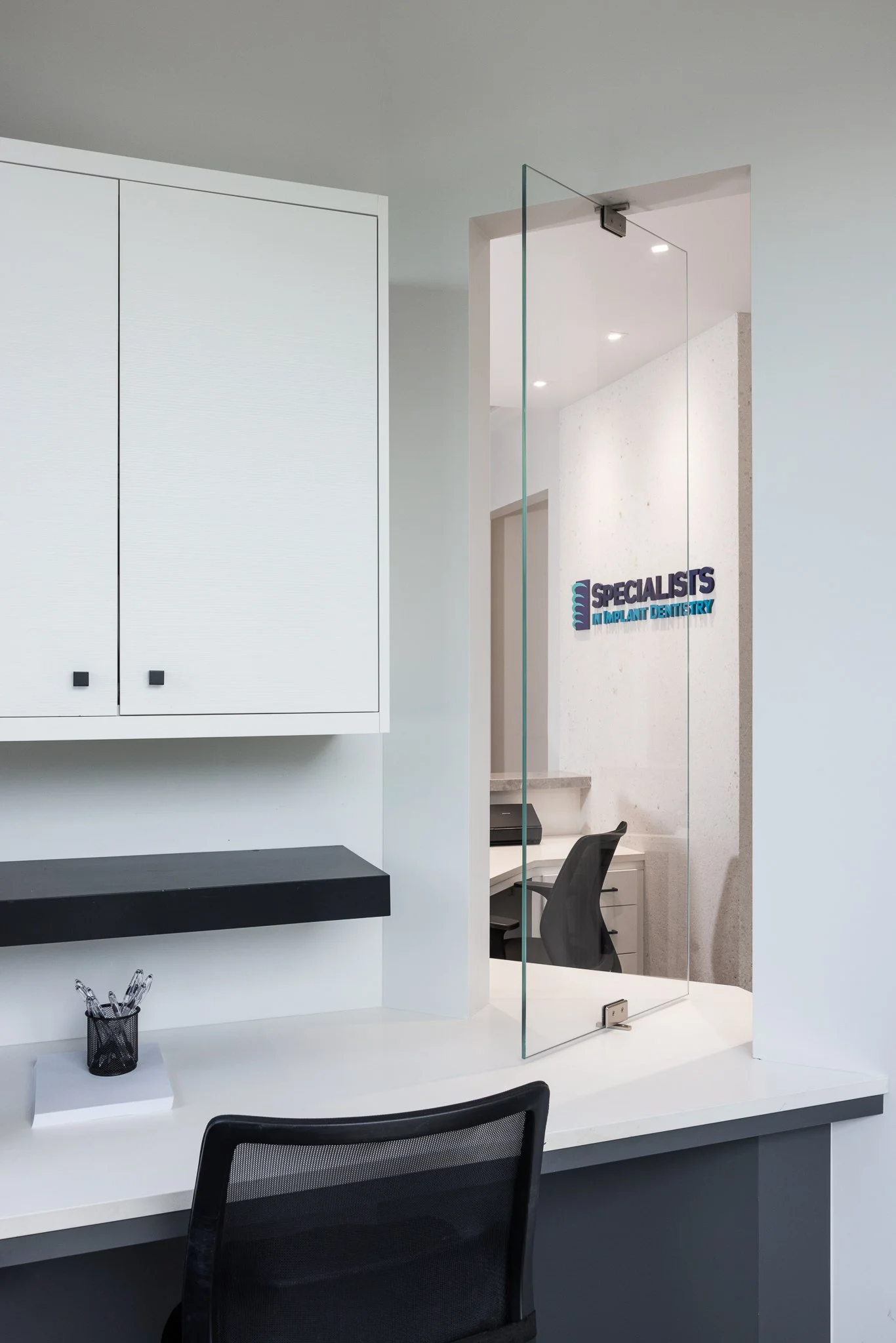 Reception area with a reception desk, a glass partition, and a wall sign that reads 'SPECIALIST'S IMPLANT DENTISTRY' in a modern dental office.