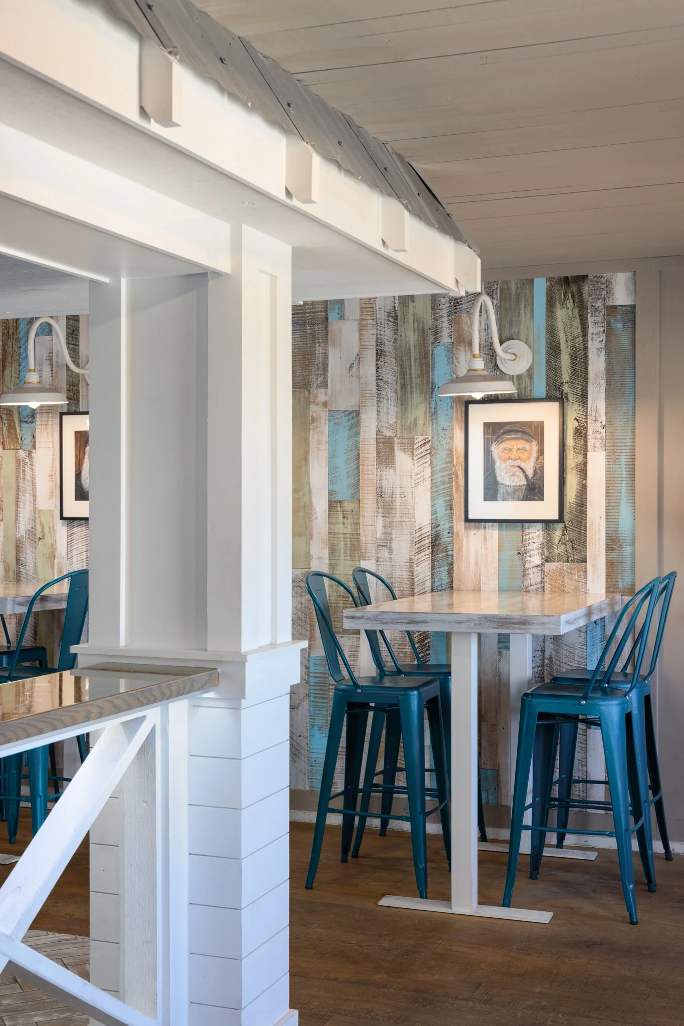 Interior of a cafe with blue chairs around a high table, a wall with rustic wood and teal accents, framed art, and wall-mounted lamps.