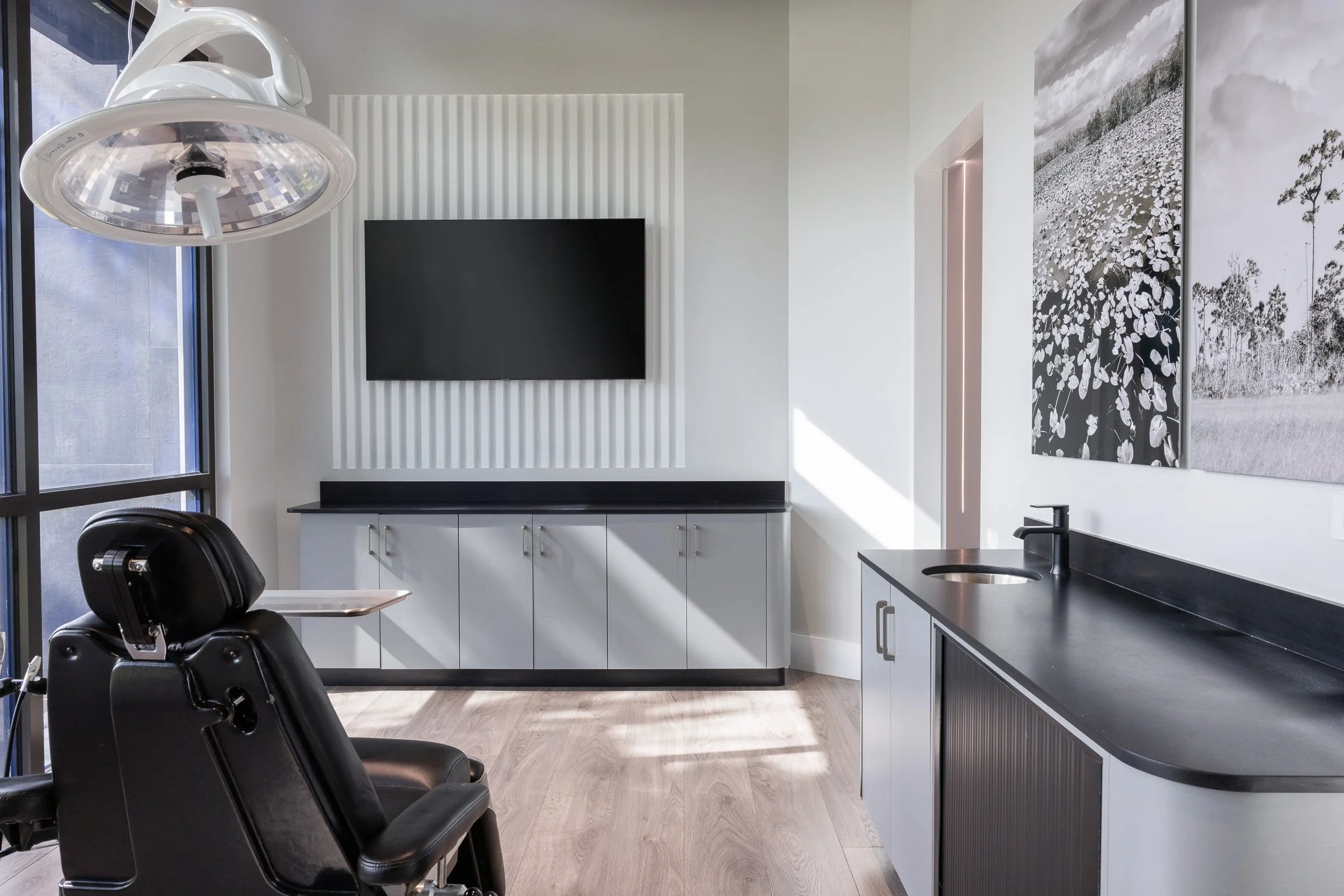 Modern dental office with black reclining chair, large window, white cabinets, black countertop, wall-mounted TV, and black-and-white artwork.