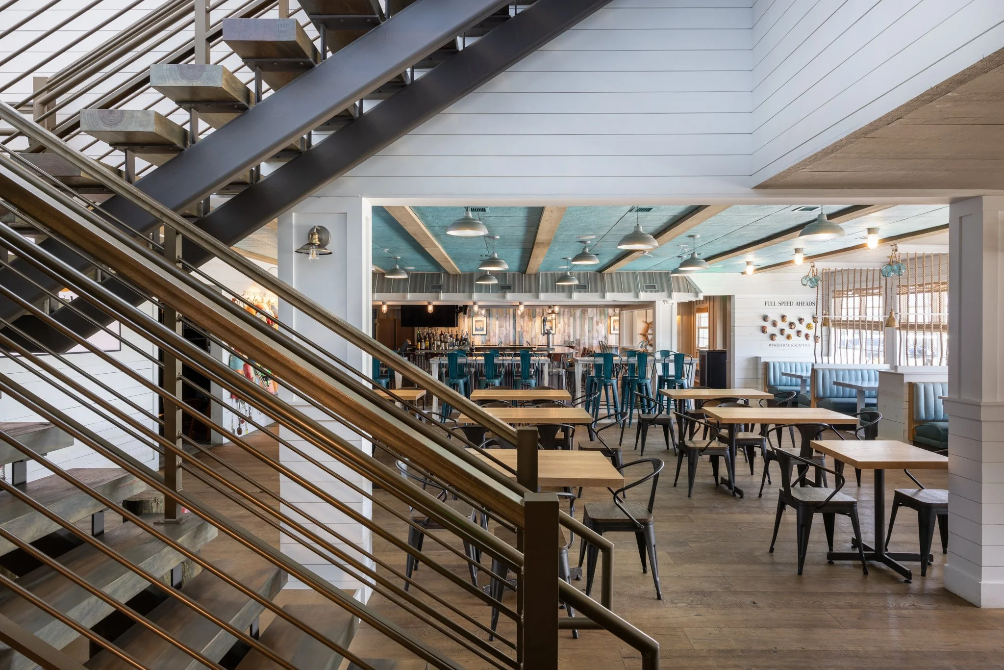 Interior view of a modern restaurant or cafe with wooden floors, multiple tables and chairs, a bar area, and ceiling lights. Stairs with metal railing are visible in the foreground.
