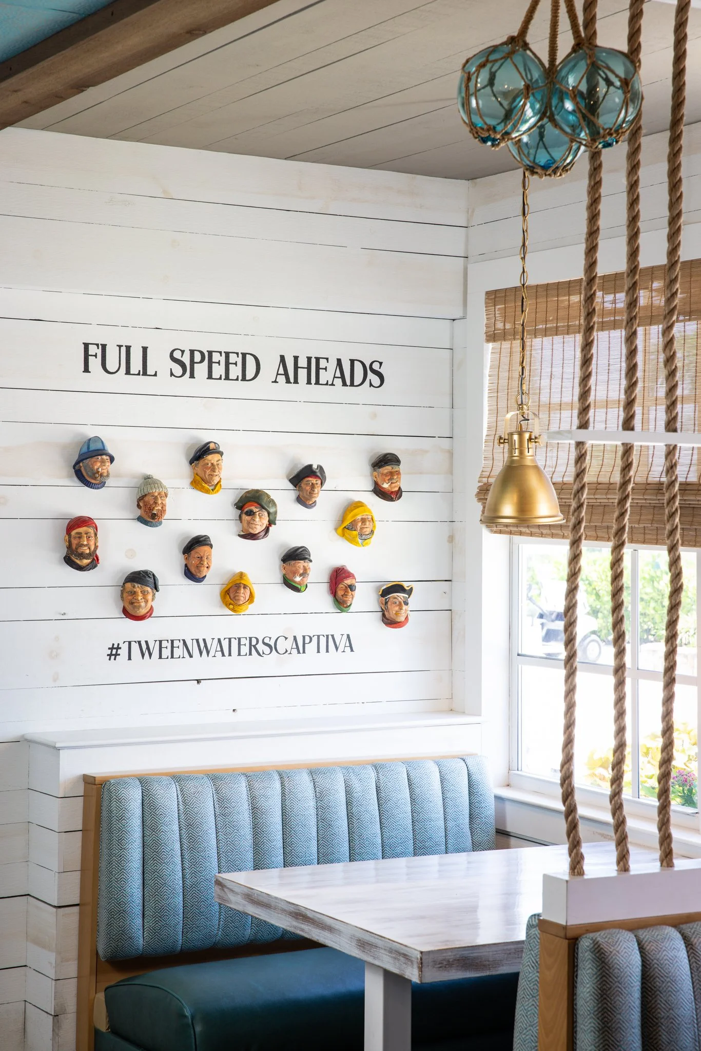 Decorative nautical themed wall with colorful carved faces of sailors with the phrase 'FULL SPEED AHEADS' and hashtag '#TWEENWATERSCAPTIVA', next to a window with bamboo blinds, hanging lights, and a booth with a wooden table.