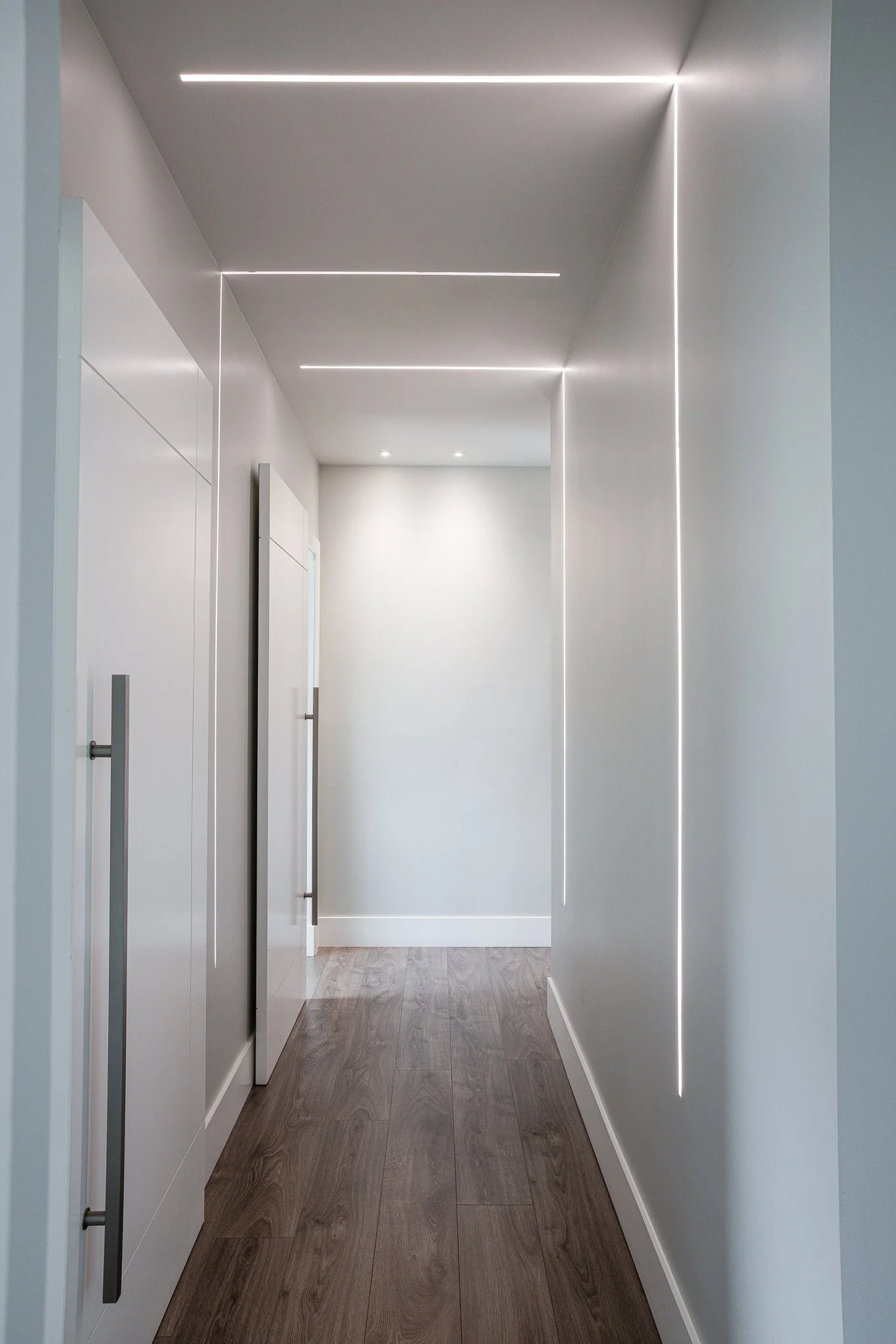 A modern hallway with white walls, gray cabinetry with long vertical handles, wood flooring, and LED lighting strips on the ceiling and wall.