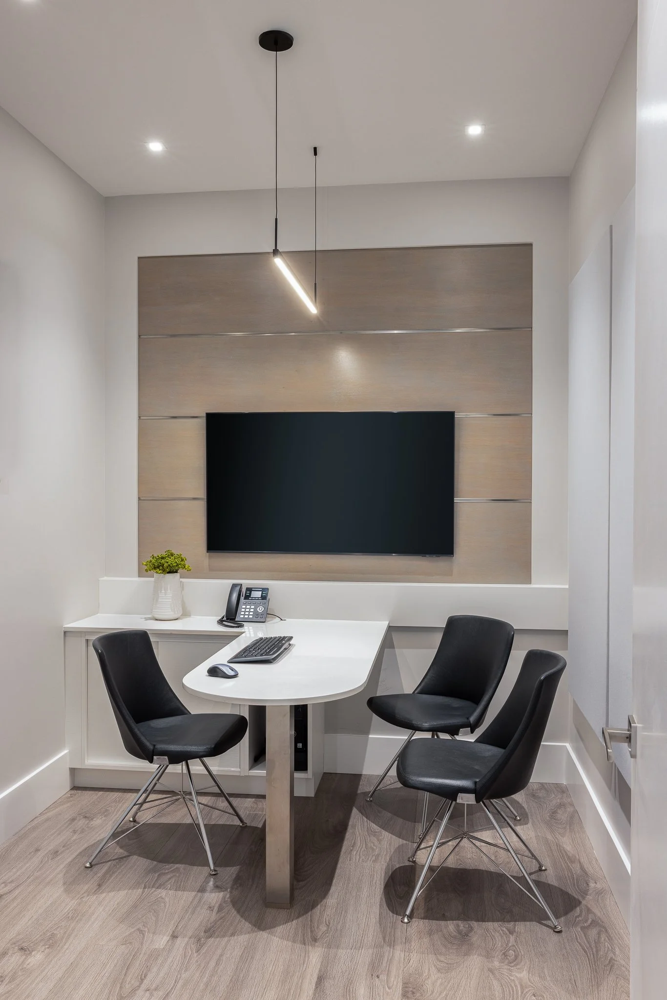 An office conference room with a white desk, four black chairs, a wall-mounted TV, a phone, a computer keyboard and mouse, a potted plant, and ceiling lights.