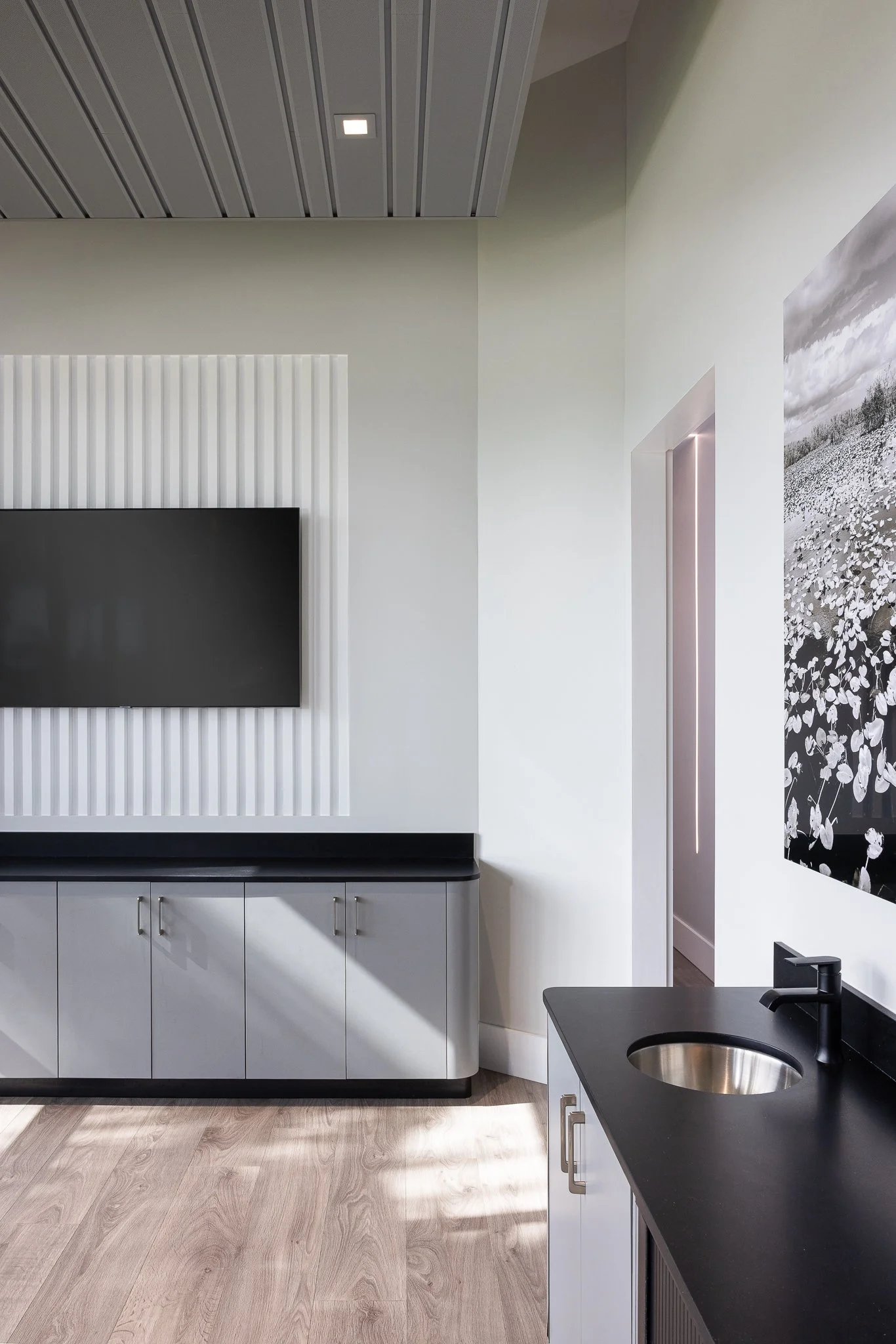 A modern interior room with a mounted flat-screen TV on a white wall, a black countertop, and a small sink with a black faucet. Light wood flooring and a white cabinet are also visible, along with a large black-and-white floral artwork on the wall.