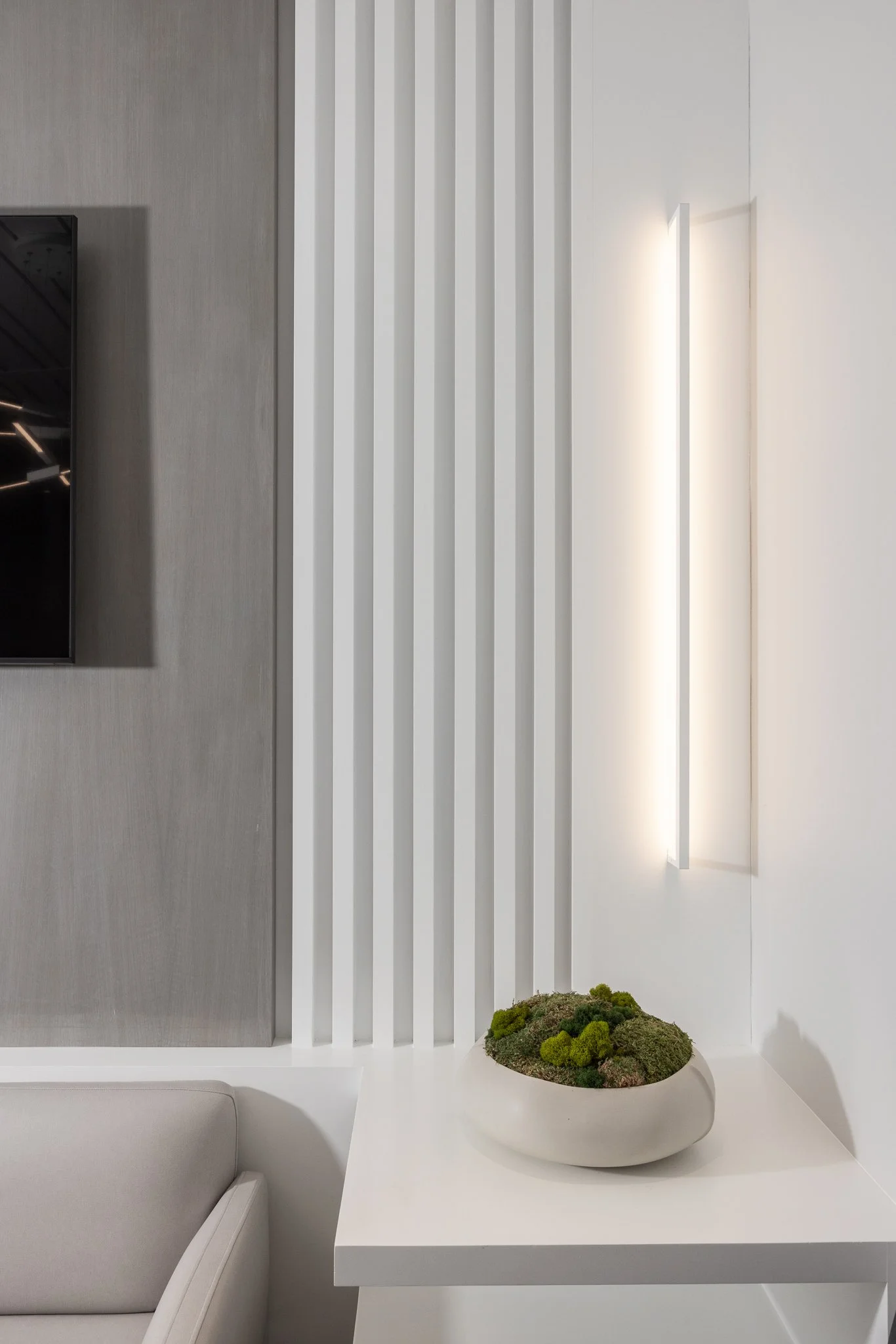 Modern interior design with a beige chair, a white side table with a decorative moss plant, and a vertical wall light on a white wall.