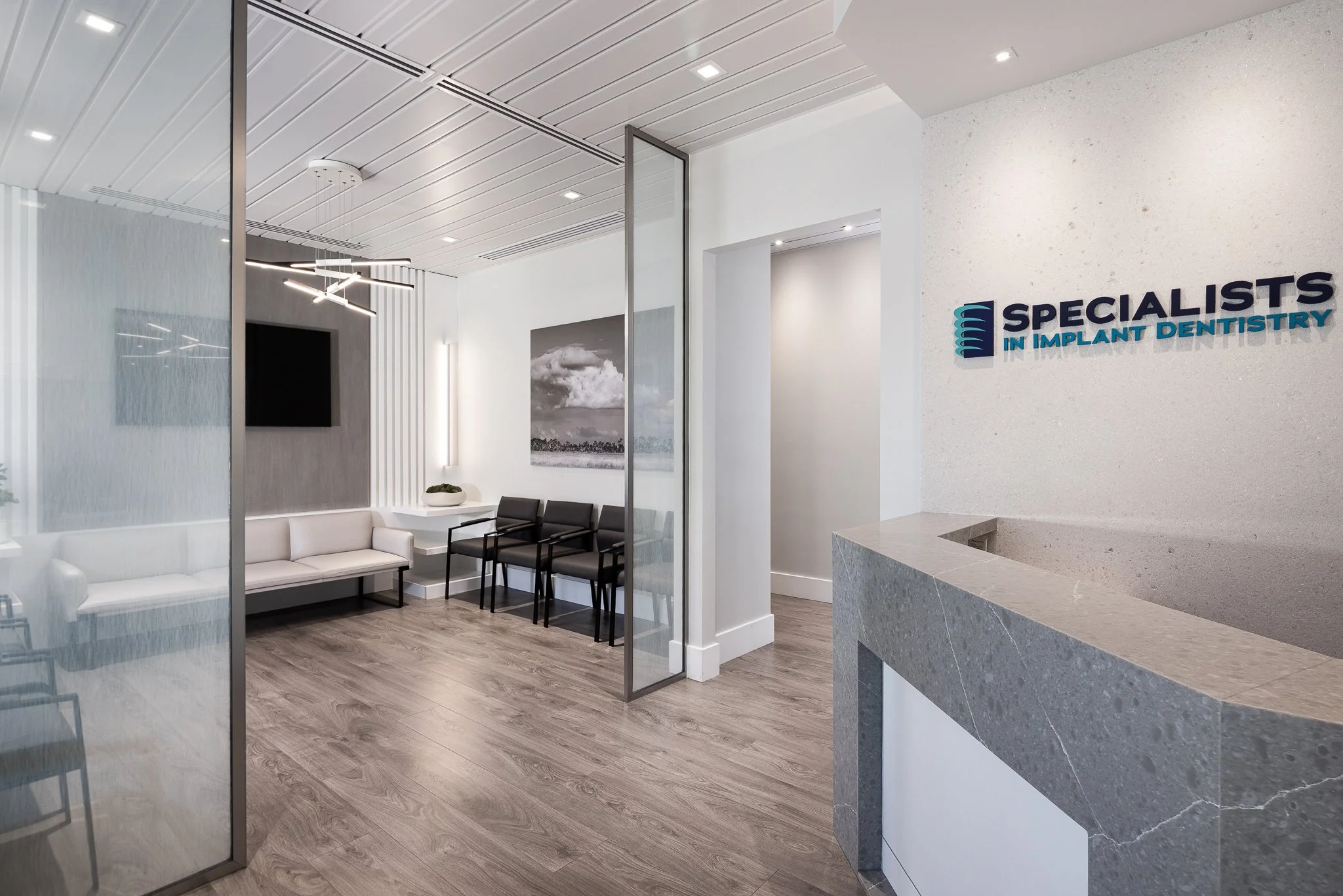 Reception area of a dental clinic with a sign that reads 'Specialists in Implant Dentistry' on the wall, featuring modern decor, a white couch, black chairs, wall art, and a flat-screen TV.