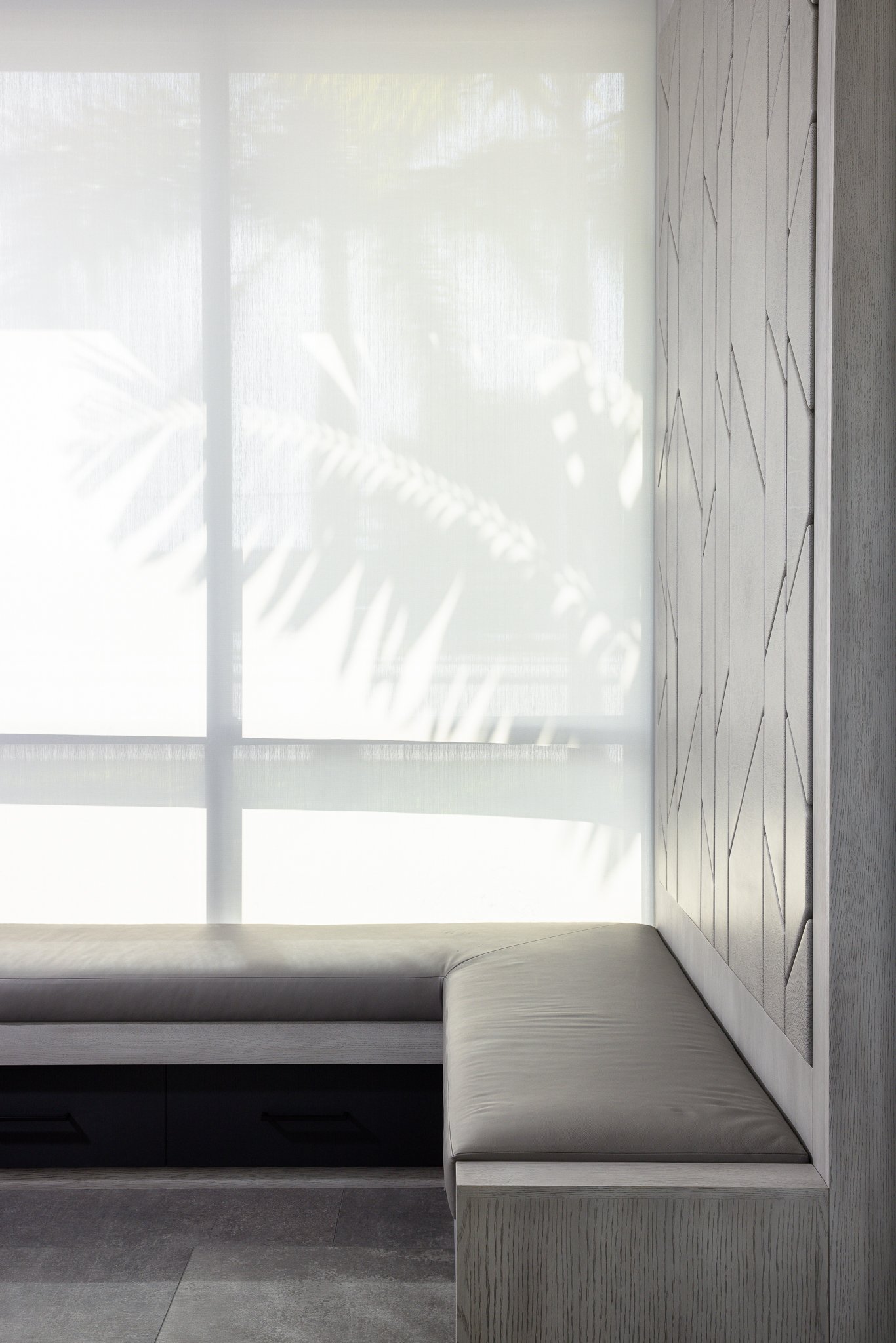 A built-in window seat with a cushion next to a large window with white sheer curtains. Shadows of leaves are cast on the curtains.