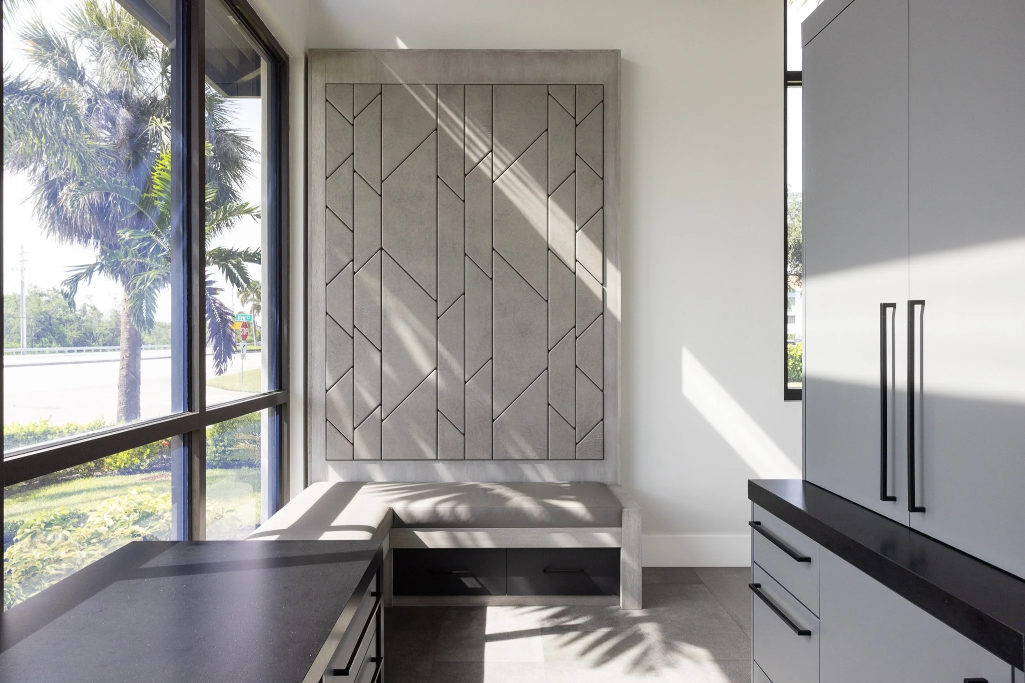 Modern kitchen corner with large window, gray panel wall with geometric pattern, white cabinets with black handles, and a built-in bench with cushion near the window.
