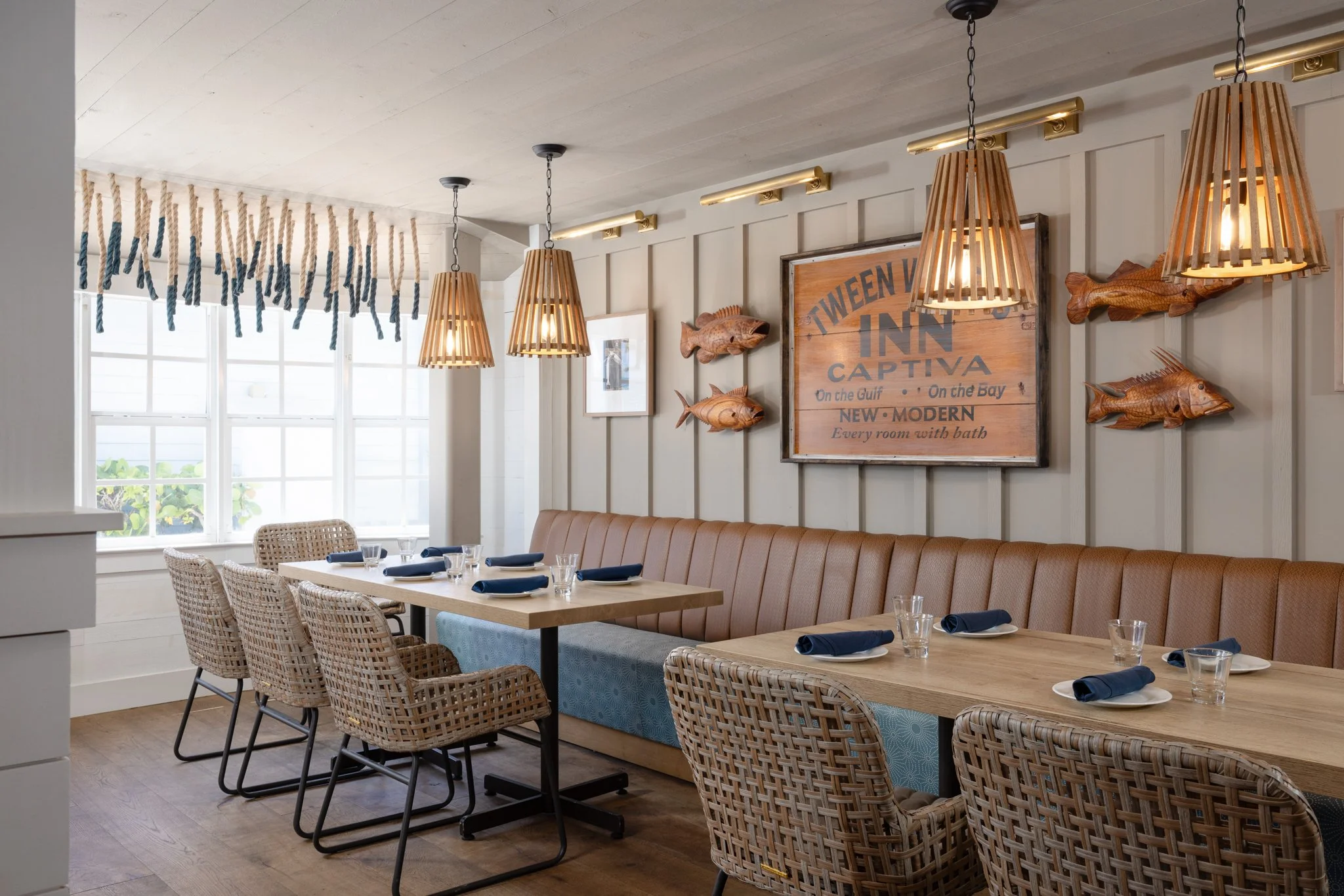 A dining area with a long wooden table set with plates, glasses, and navy napkins. Beige wicker chairs and a tan leather bench surround the table. The wall behind has wood fish decorations and a sign reading "Tween Waters Inn Captiva". Wooden pendant