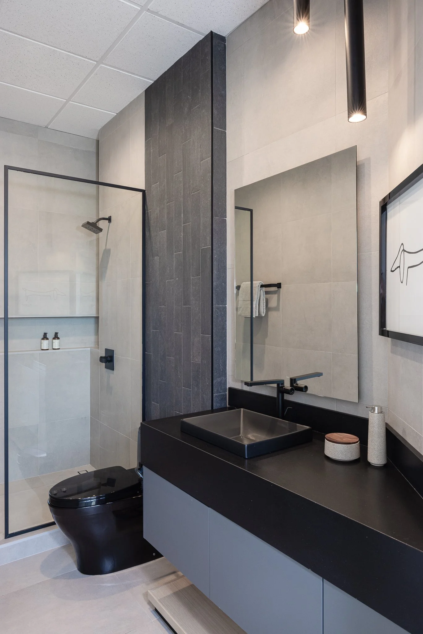 Modern bathroom with a black toilet, black sink, and large mirror. There is a shower area with a glass door and a wall-mounted showerhead. The decor features light-colored tiles with a dark accent wall and black fixtures.