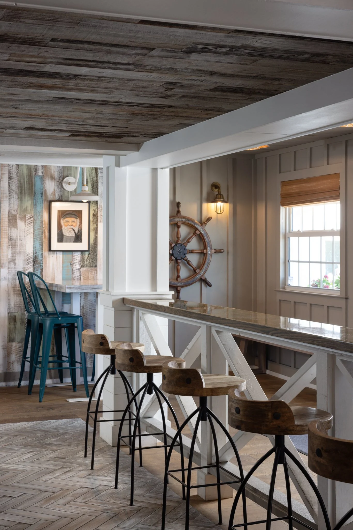 Interior of a rustic style cafe or bar with wooden bar stools, a high counter, nautical decor including a ship's wheel, and a framed portrait on wood-paneled wall.