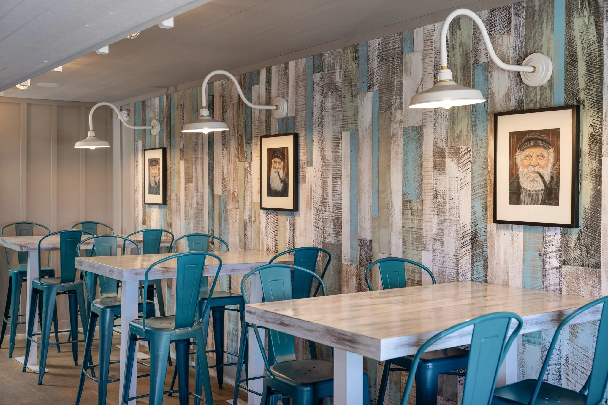 Interior of a cafe with wooden tables, teal metal chairs, a rustic wood wall with framed portraits, and white industrial-style lamps.