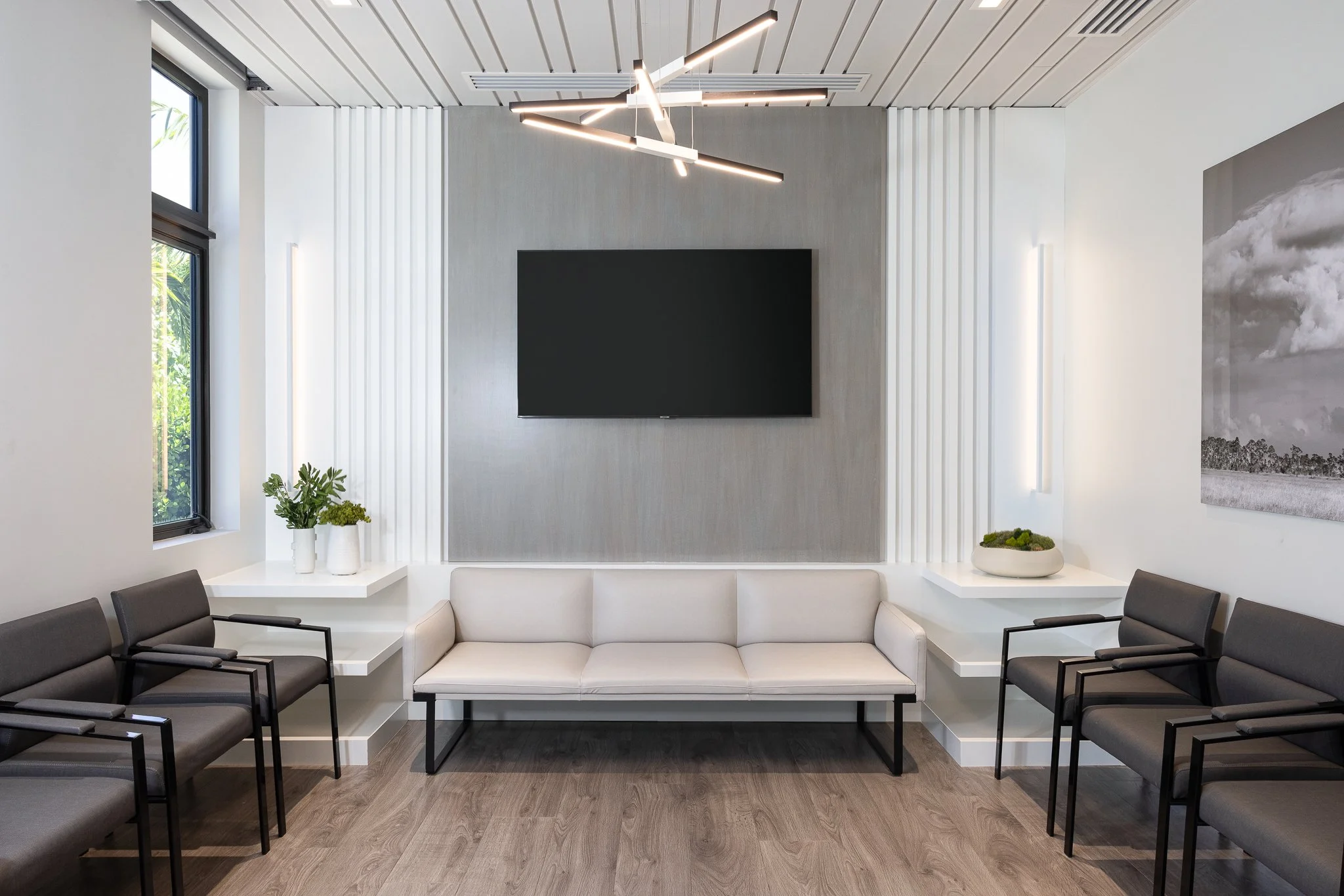 A modern waiting room with a white couch, six dark gray chairs, a large black wall-mounted TV, and a contemporary light fixture on the ceiling. Two windows with plants on white shelves, and black-and-white artwork on the walls.