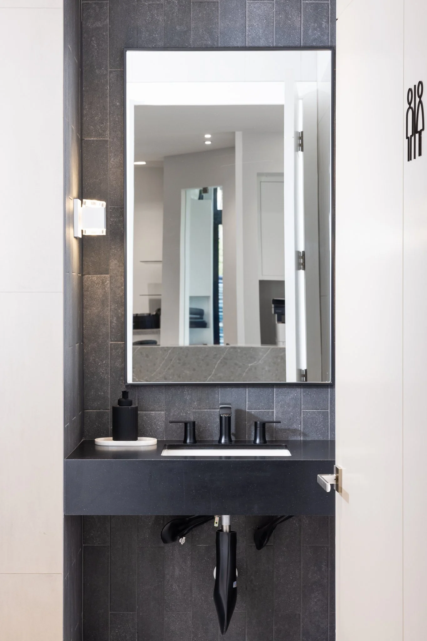 Modern bathroom with a black framed mirror above a black sink, dark gray tiled wall, white walls, wall-mounted light fixture, and black accessories.