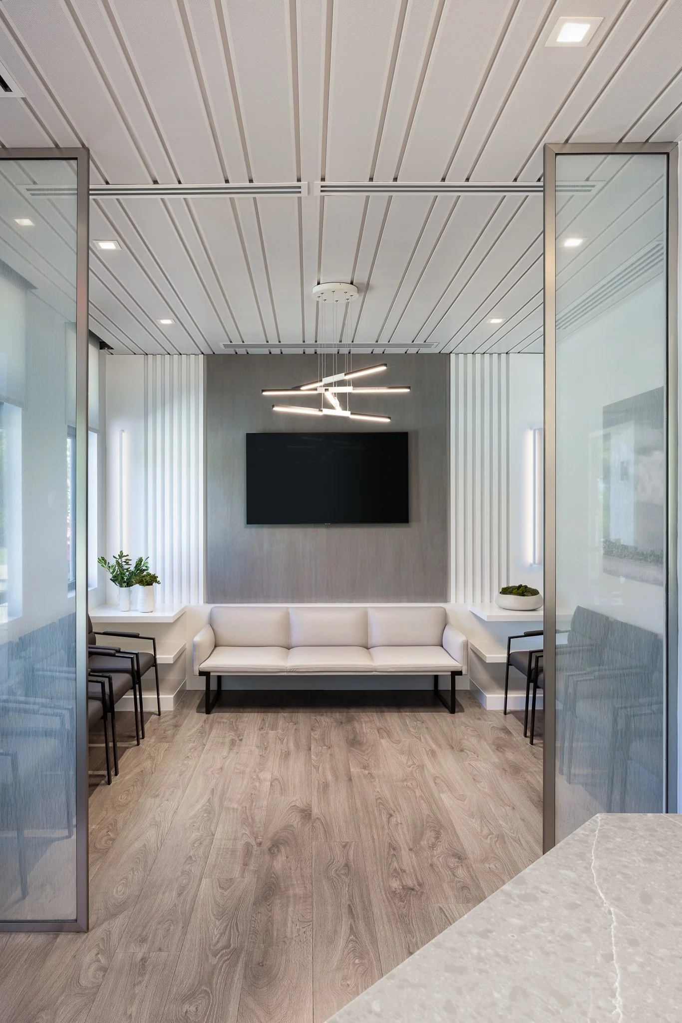 Modern waiting room with white sofa, black chairs, potted plants, large TV, gray accent wall, and contemporary ceiling light fixture.