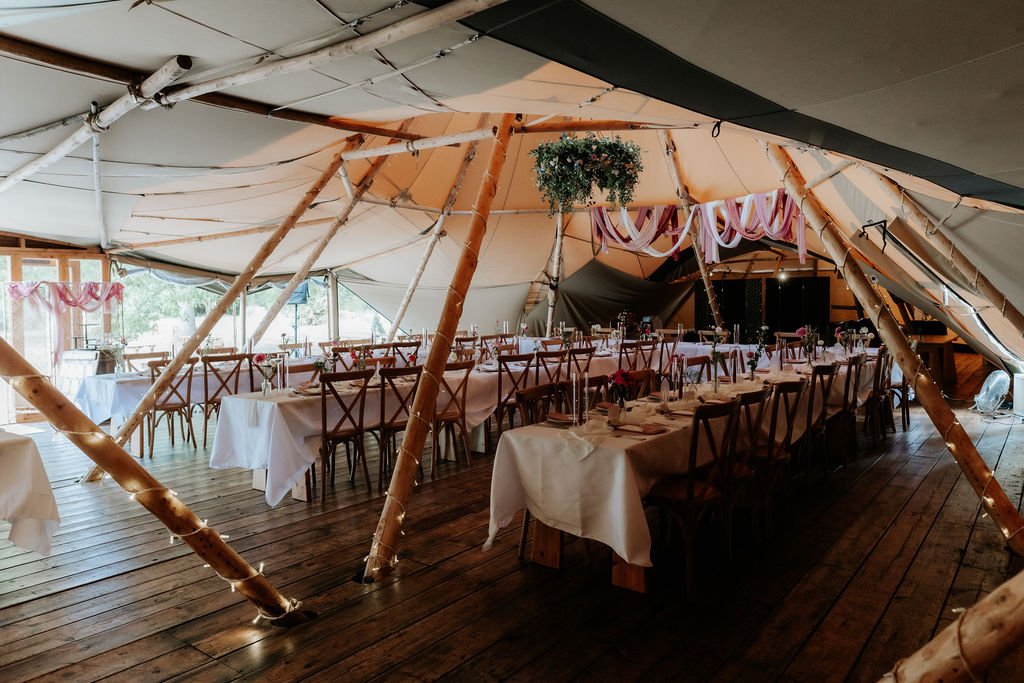 Wide view of the wedding reception inside Tipis at Whatton House, with wooden dining tables, festoon lights and a hanging floral halo.