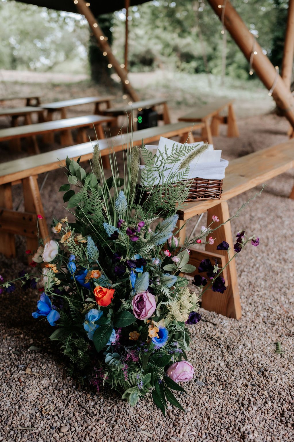 Artificial wedding aisle flowers in blue, pink, orange and purple with mixed greenery beside wooden chairs at Whatton House.