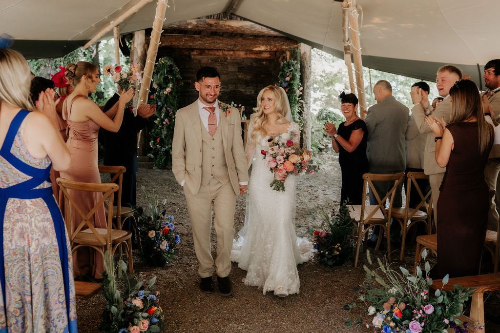 Newly married couple walking back down the flower-lined outdoor ceremony aisle at Tipis at Whatton House.