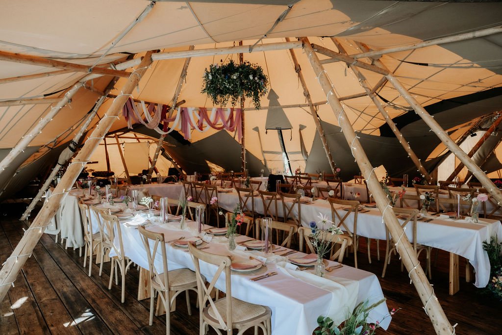 Wedding breakfast tables inside Tipis at Whatton House, with a hanging floral halo, pastel drapes and colourful table flowers.