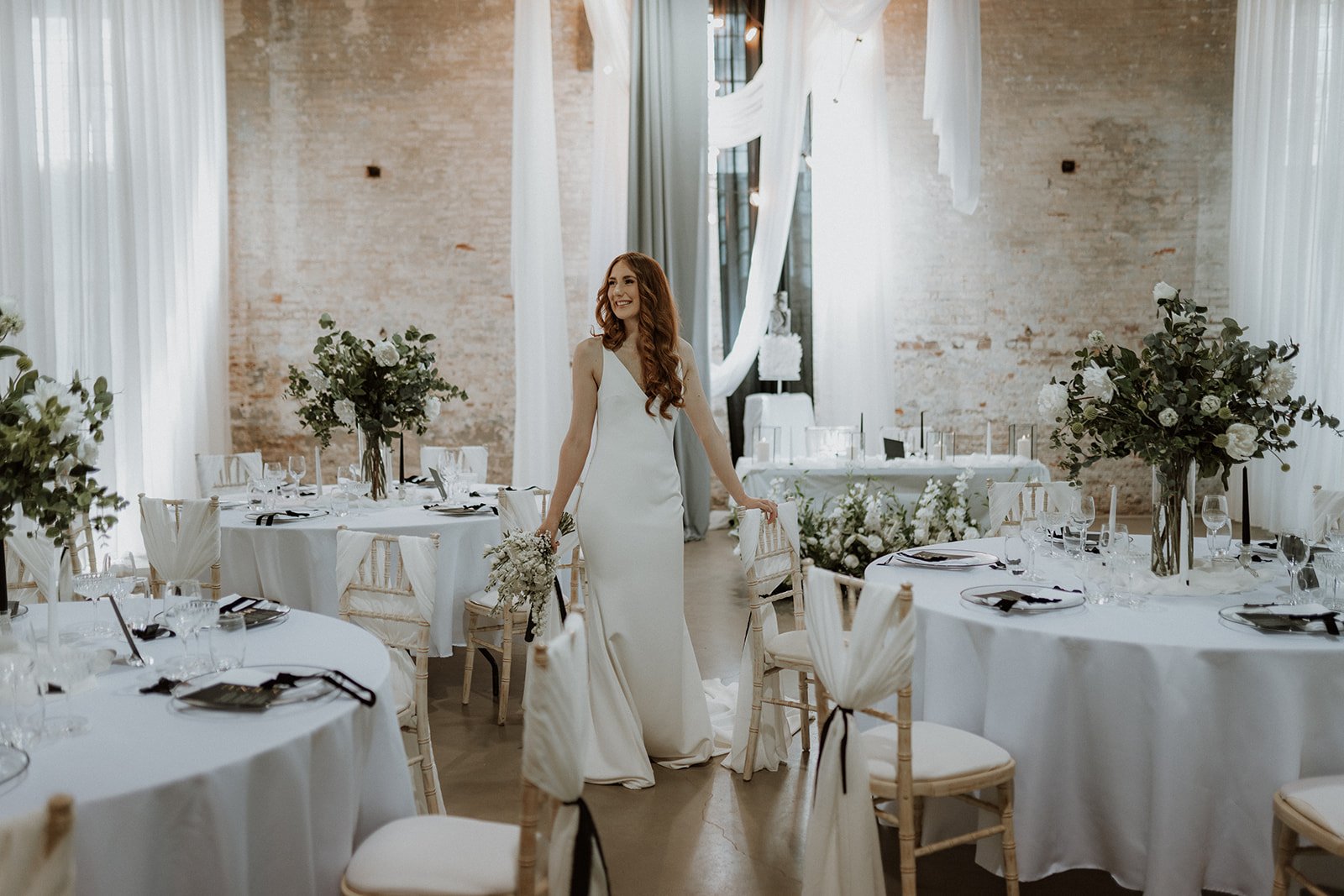 Bride standing in a wedding reception at The Riding School Calke Abbey styled with white artificial flowers, greenery and table centrepieces